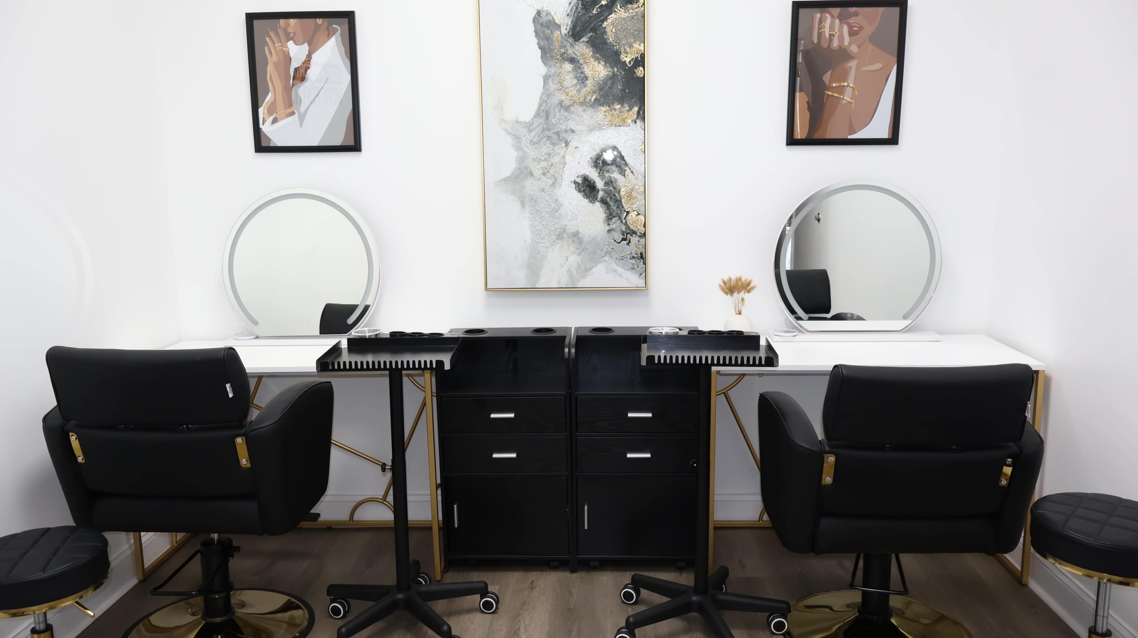 The image shows a modern beauty studio with two black salon chairs, two round mirrors, and a central black storage unit, accented by framed artworks on the walls.