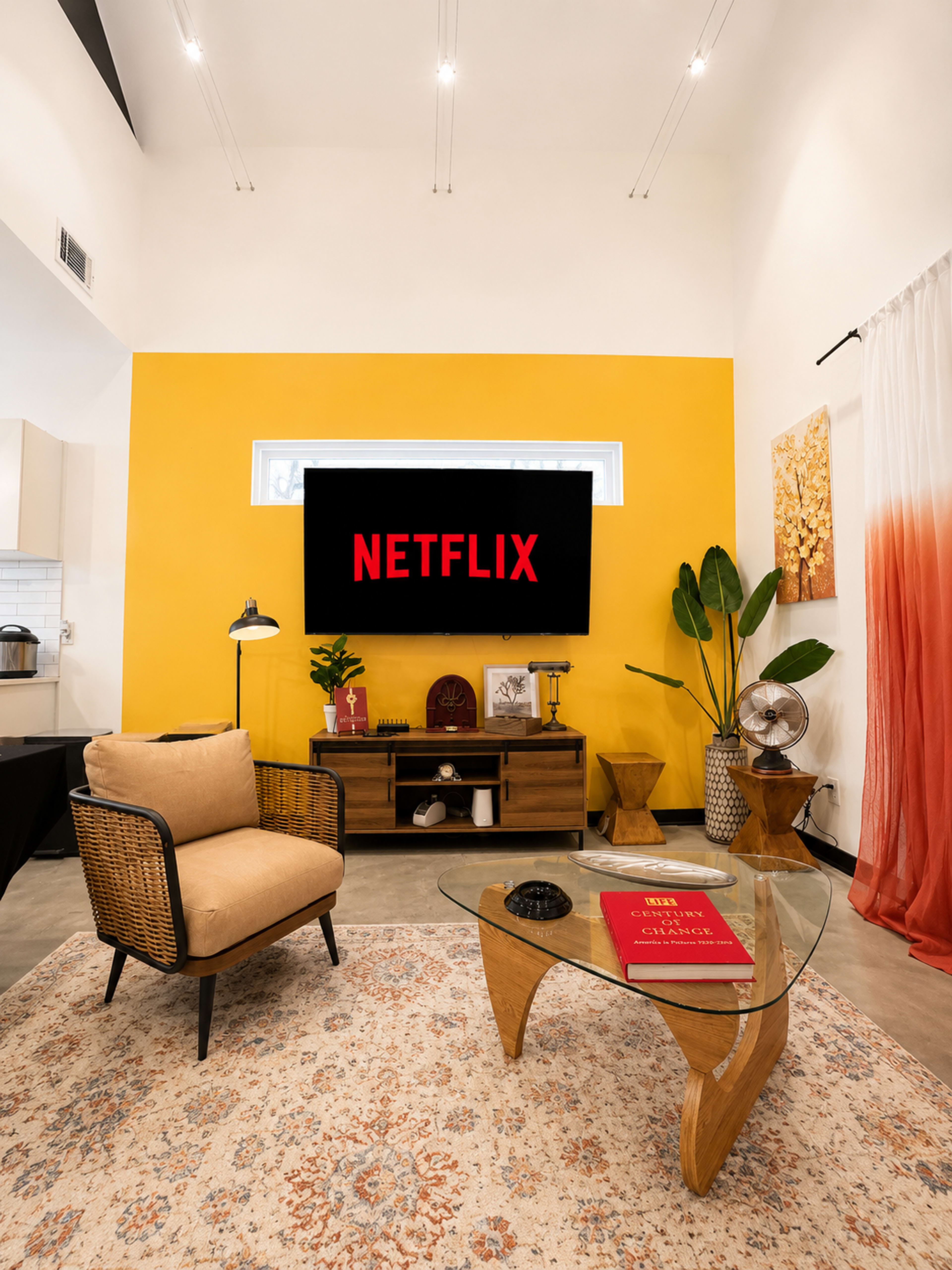 The image shows a modern living room with a yellow accent wall, featuring a television displaying the Netflix logo, a brown armchair, a glass coffee table, and decorative plants.