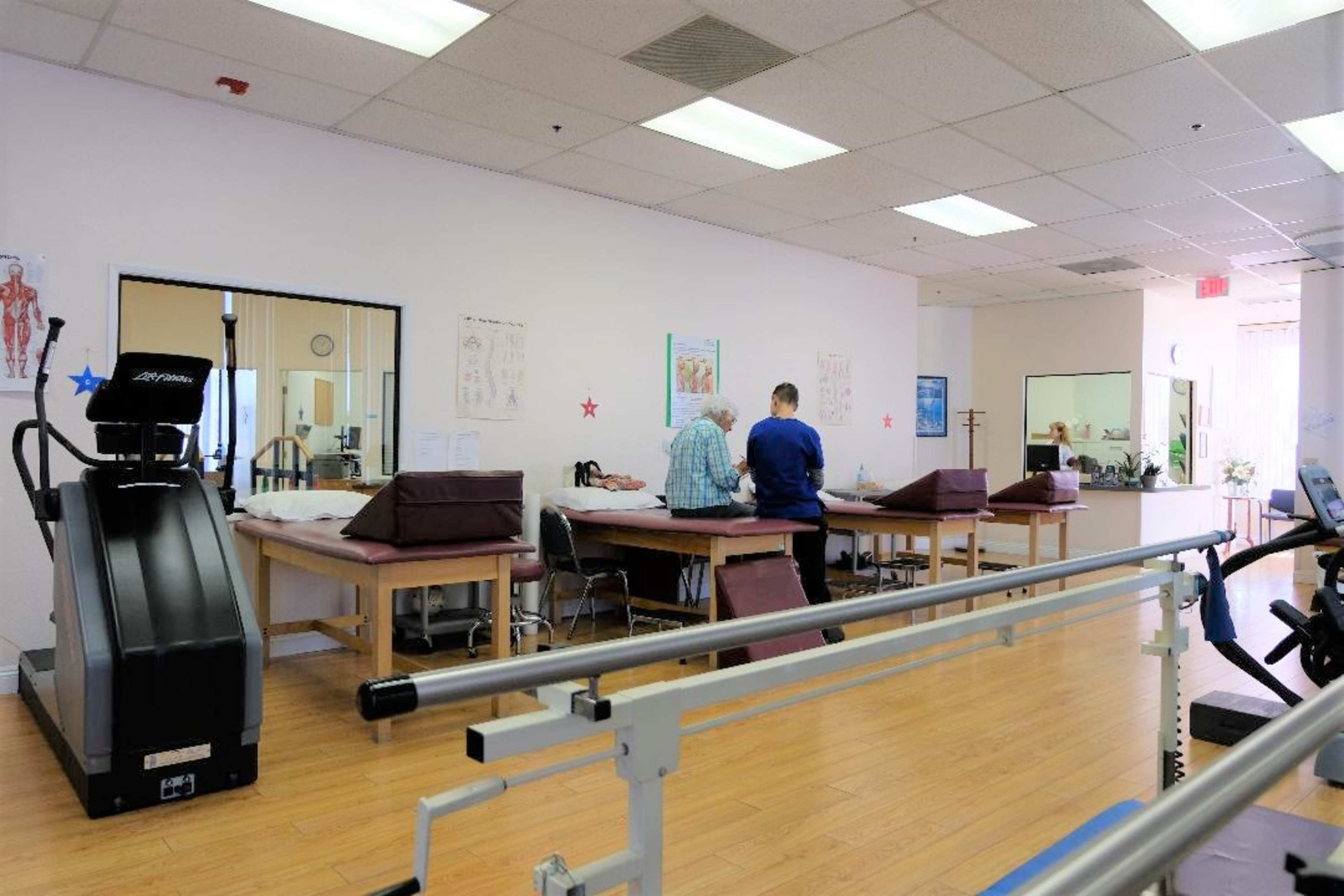 A physical therapy clinic with treatment tables, a treadmill, and patients receiving care from therapists.