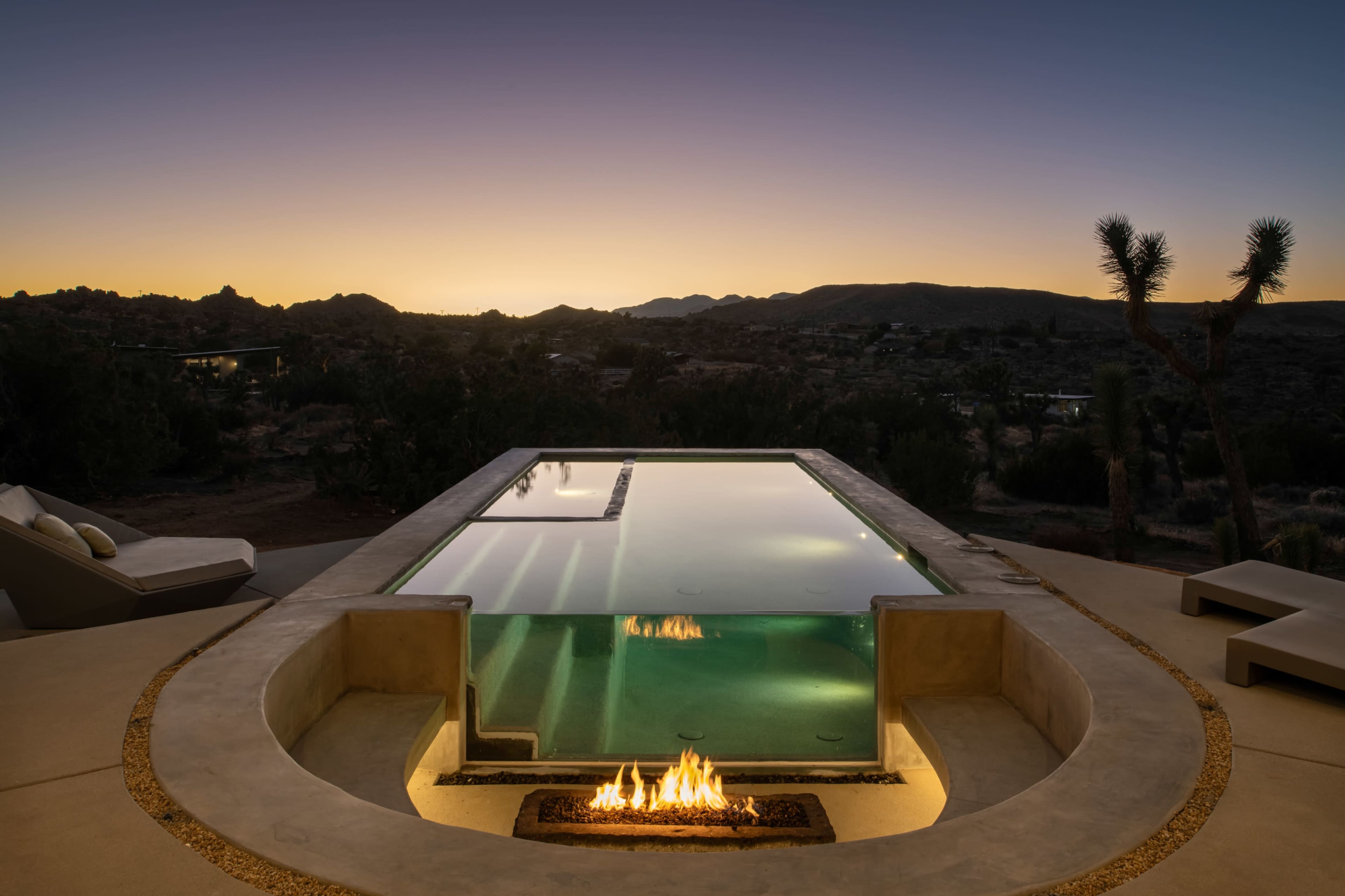 The image shows a sleek infinity pool with a fire feature surrounded by desert landscape during twilight.