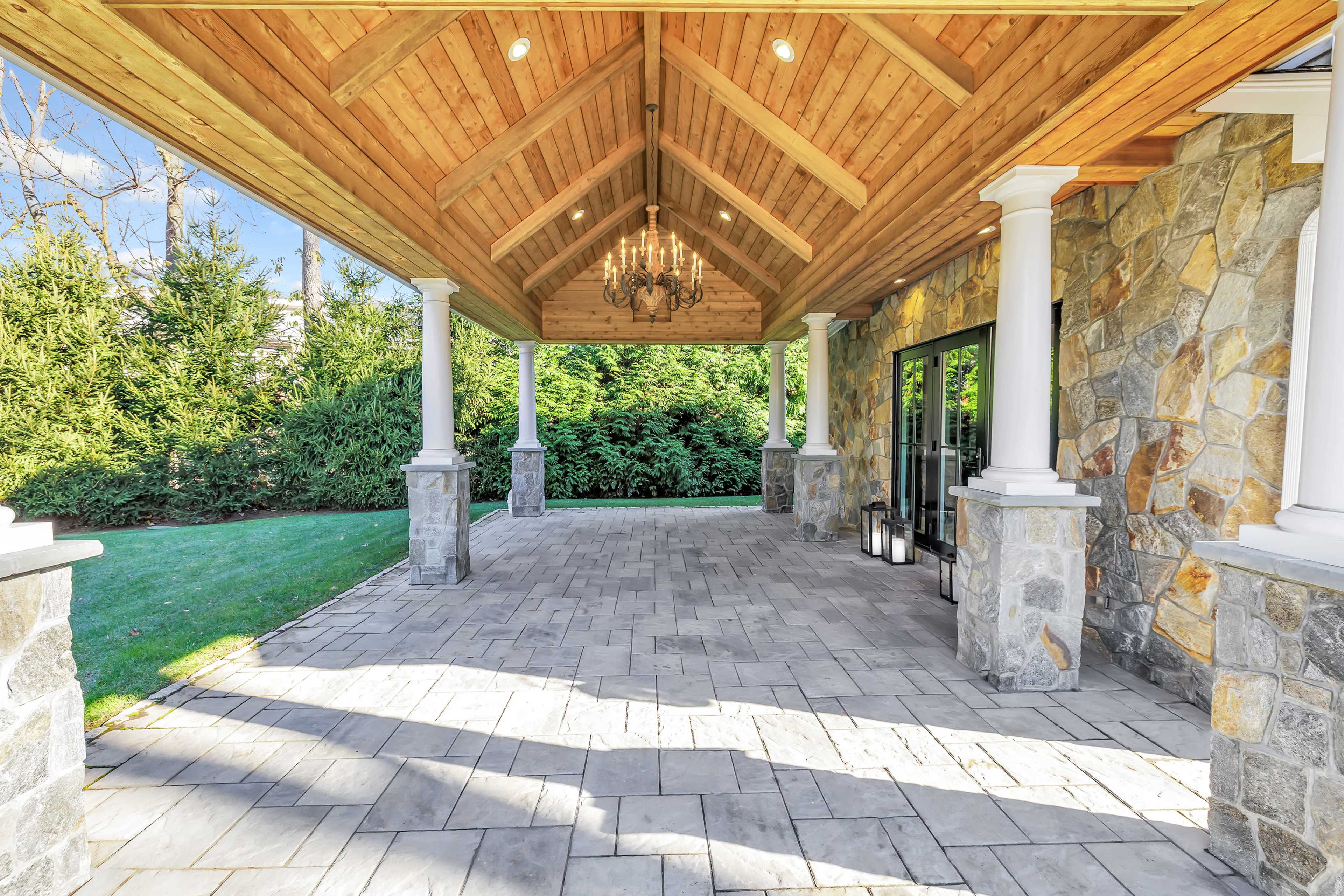 The image shows a spacious patio area with a stone floor, wooden ceiling beams, and elegant columns, surrounded by greenery.