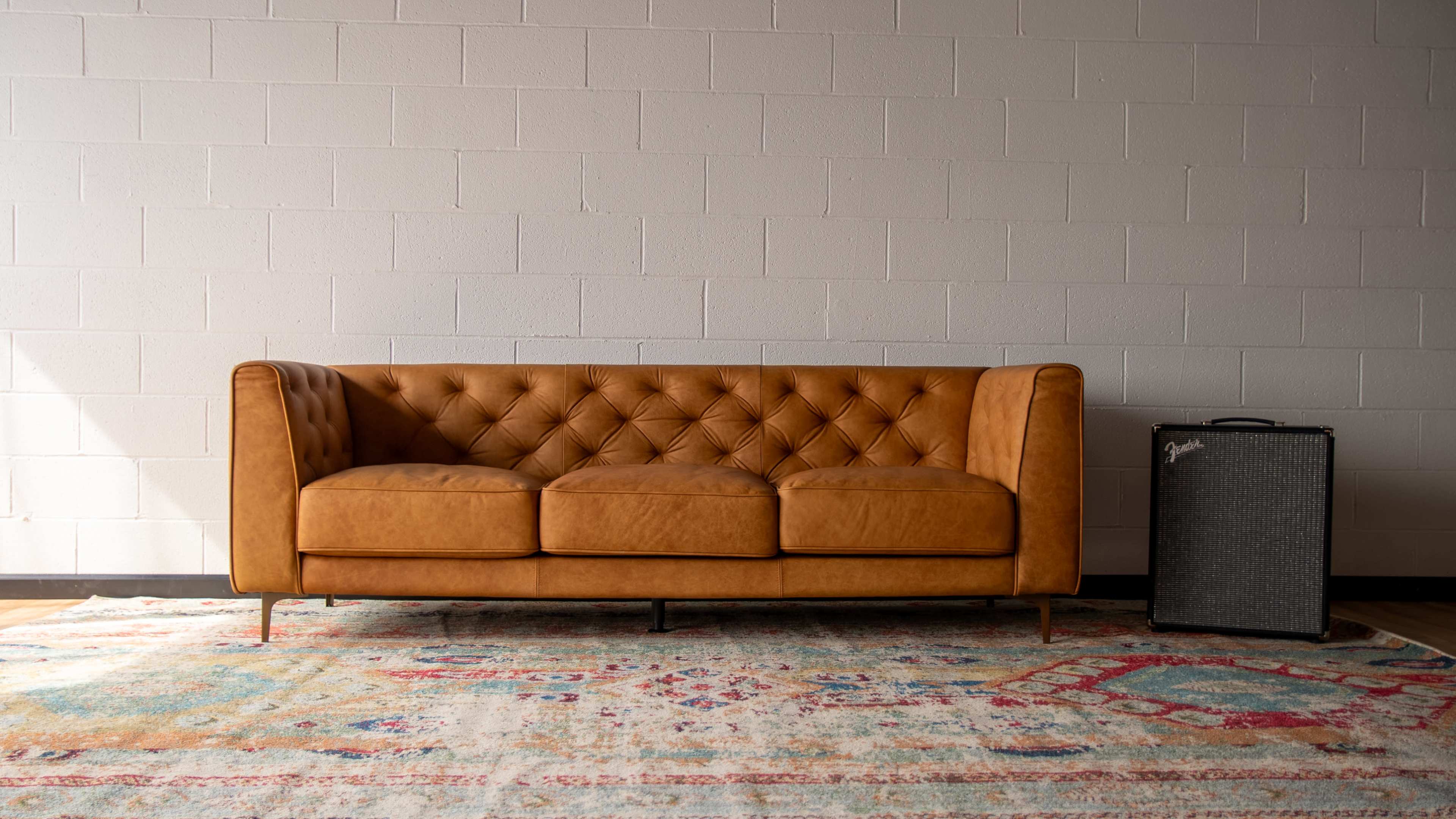 A tan leather sofa with tufted cushions is positioned in front of a textured area rug and a black amplifier against a white brick wall.
