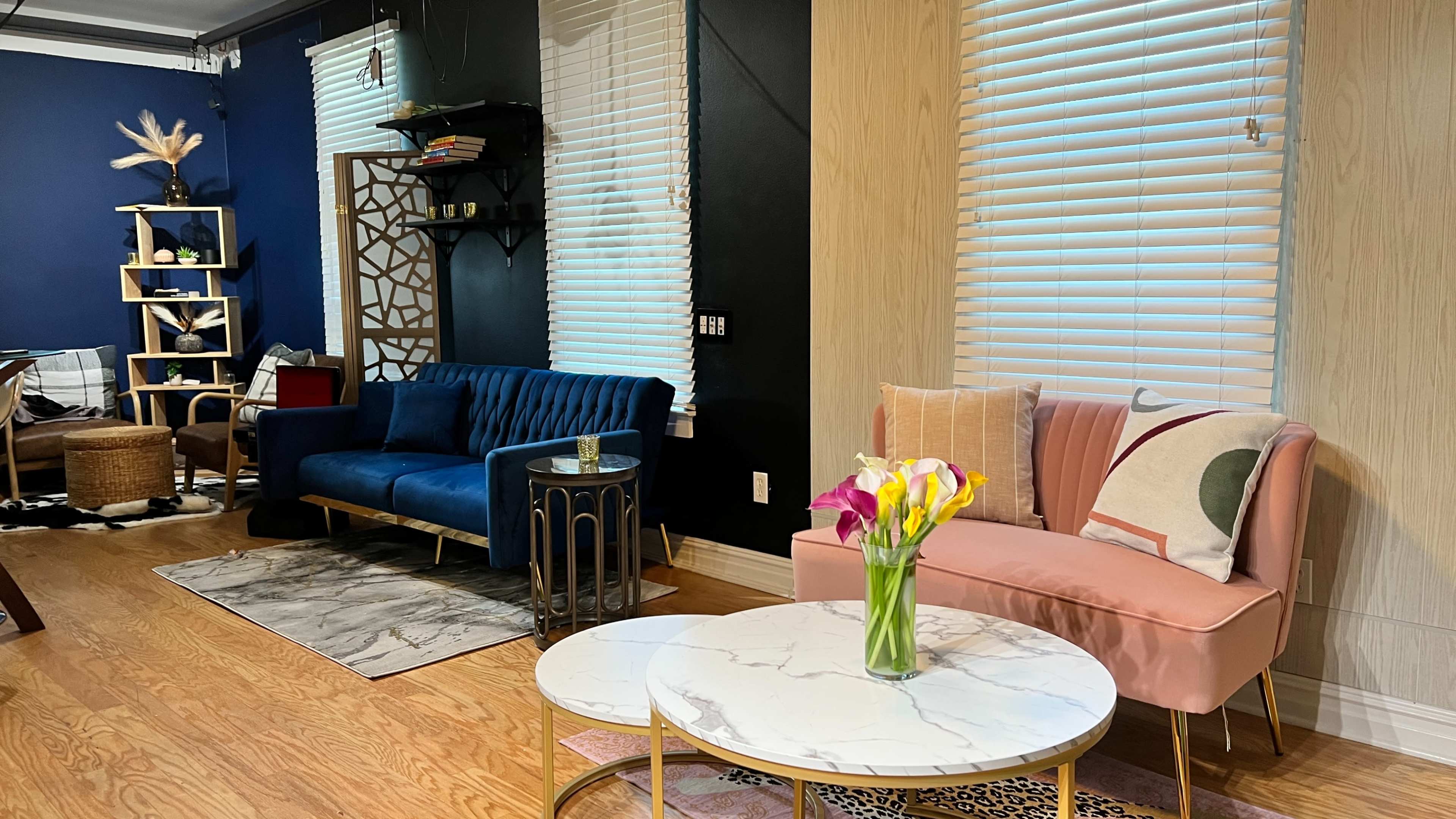 The image shows a modern living room with a blue velvet sofa, a pink armchair, and a round marble coffee table, accented by a vase of fresh flowers.