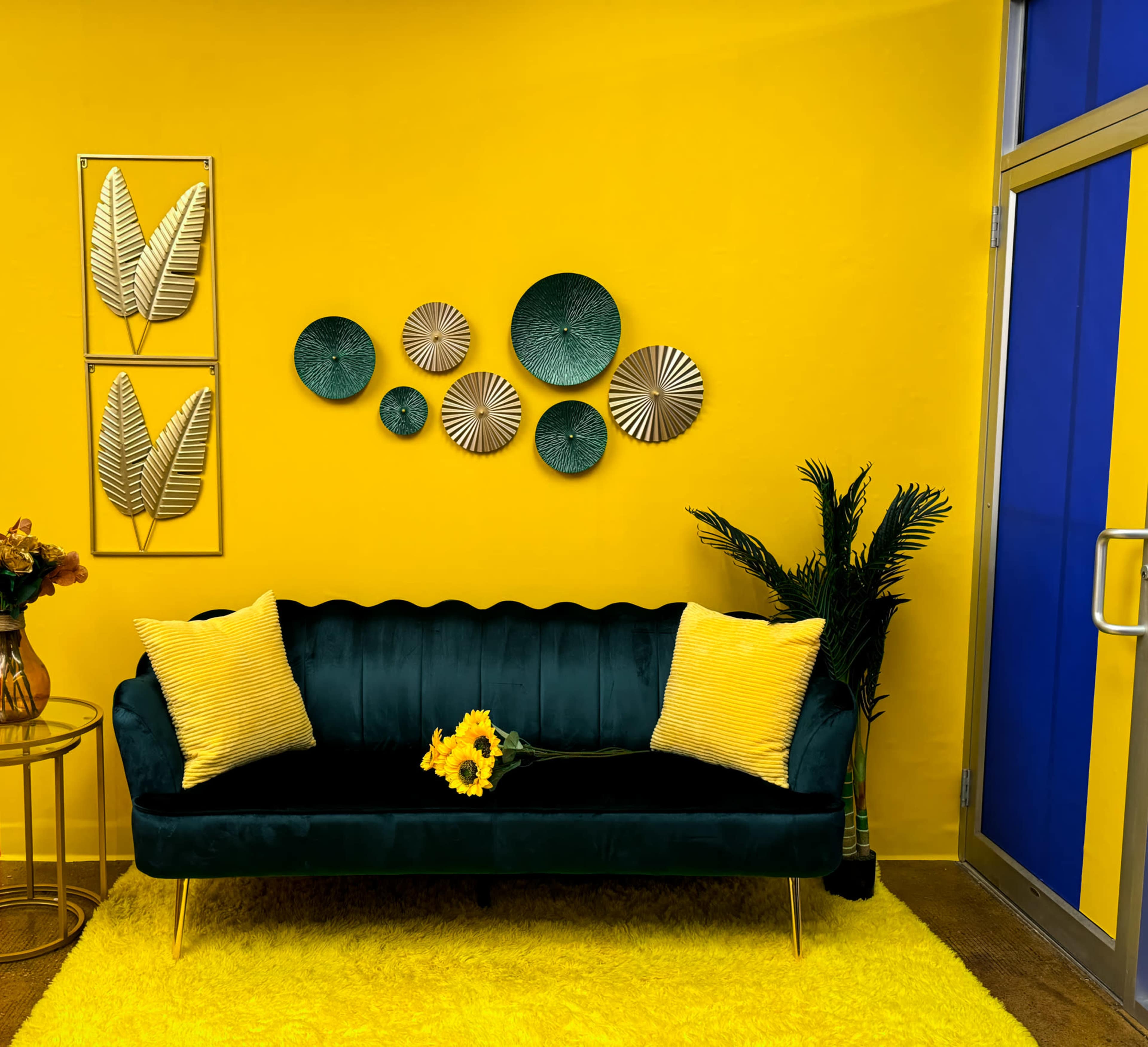 A green velvet sofa with yellow pillows is placed against a bright yellow wall, decorated with circular wall art, next to a small table with a vase of flowers and a potted plant.