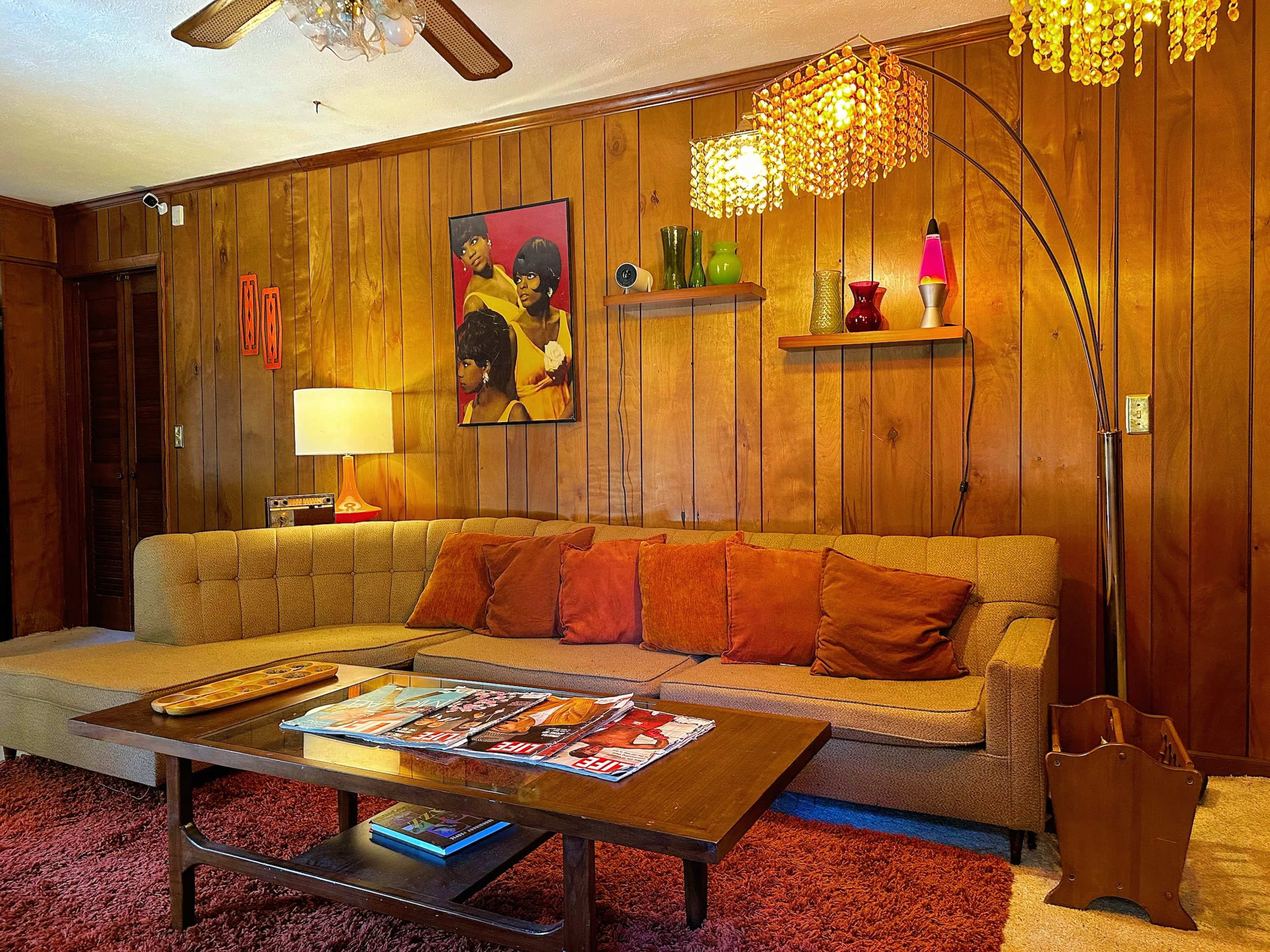 The image shows a mid-century modern living room with a brown wood-paneled wall, a beige sofa with orange cushions, and a coffee table displaying magazines.