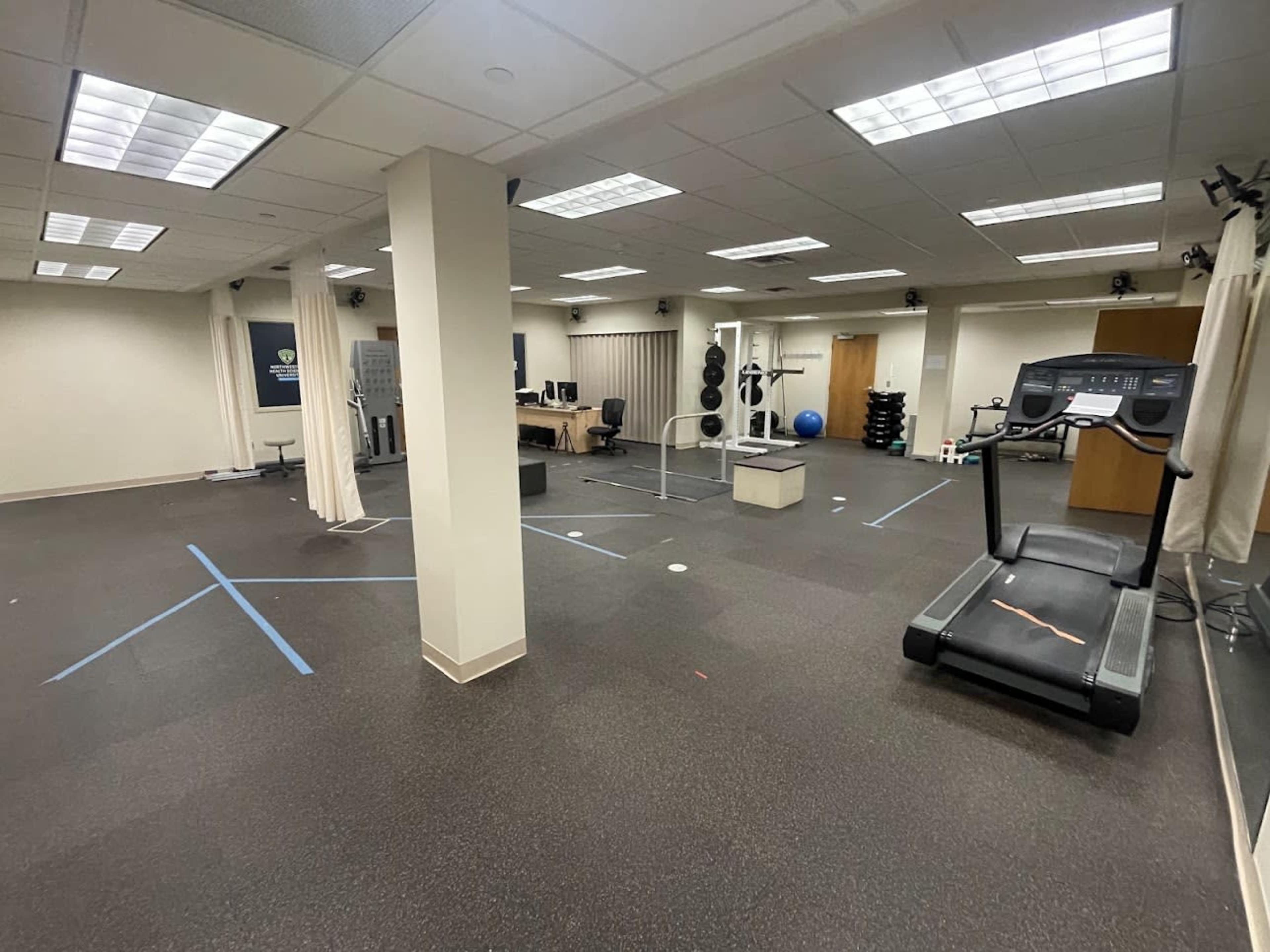 A sparsely equipped gym with exercise machines, weights, and marked areas on the floor.