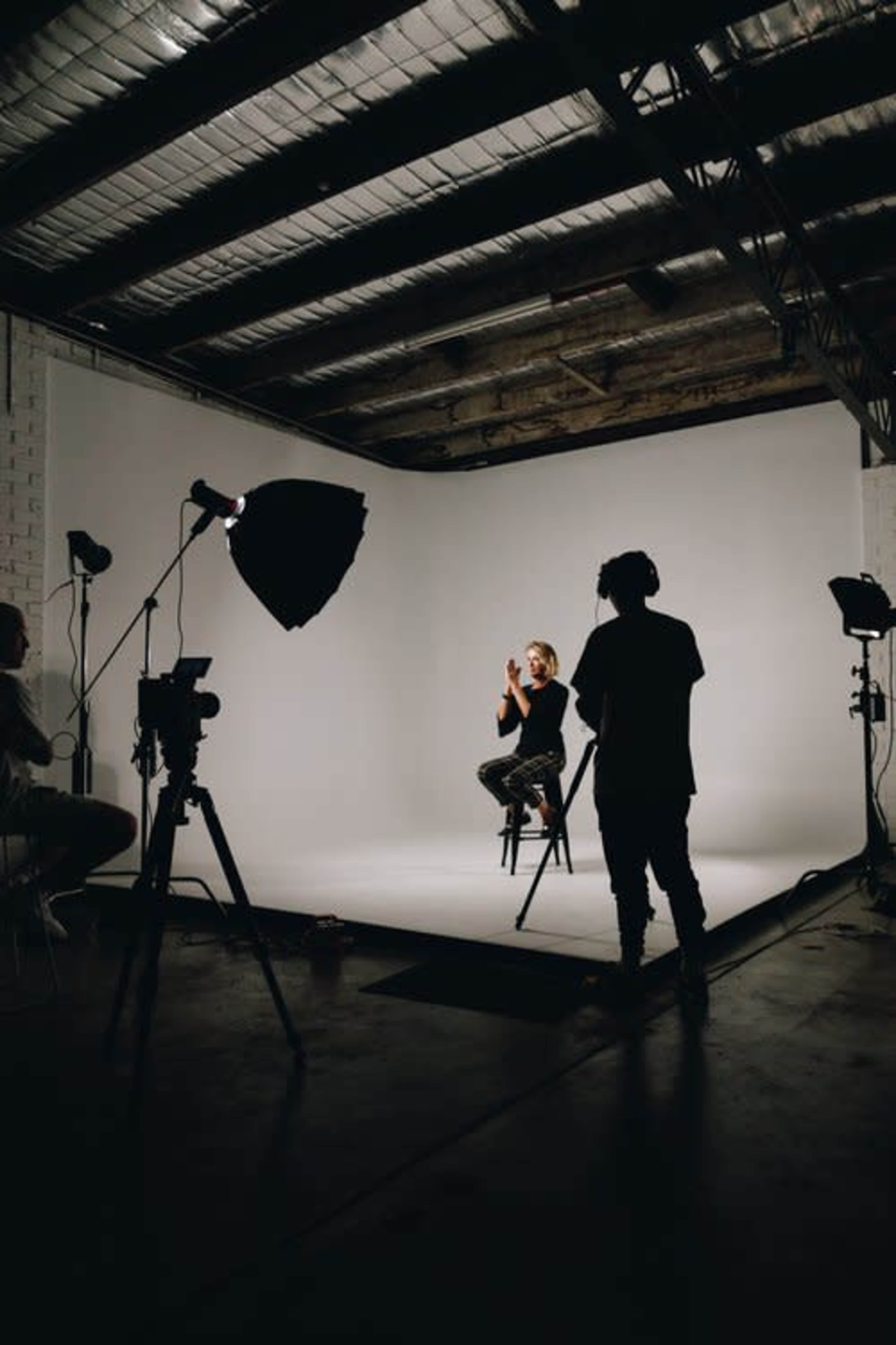 A person sits on a stool in a studio setting, illuminated by multiple lights and surrounded by cameras on tripods.