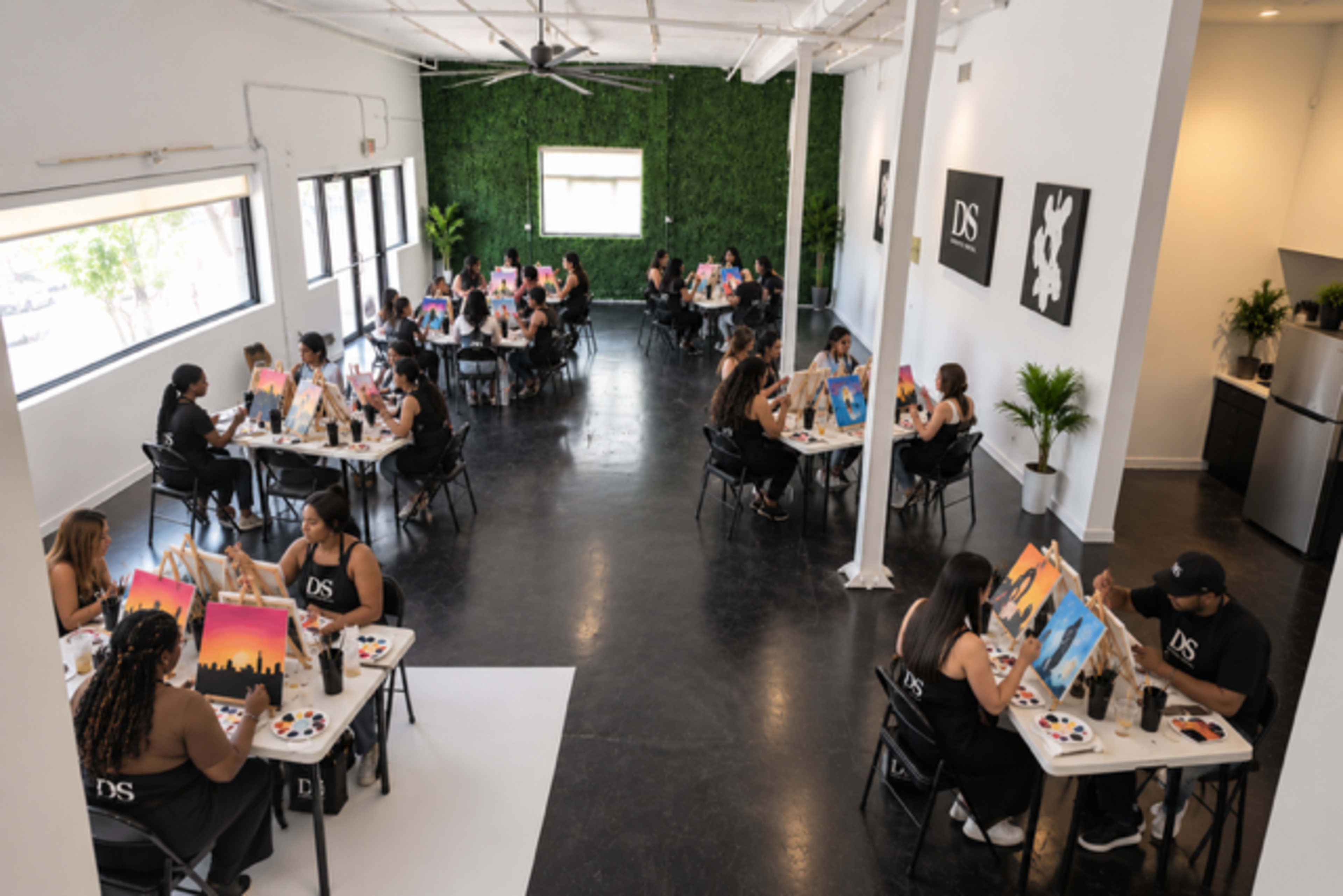 The image shows a spacious art studio with participants seated at tables, each painting on canvases in a well-lit environment with green wall decor.