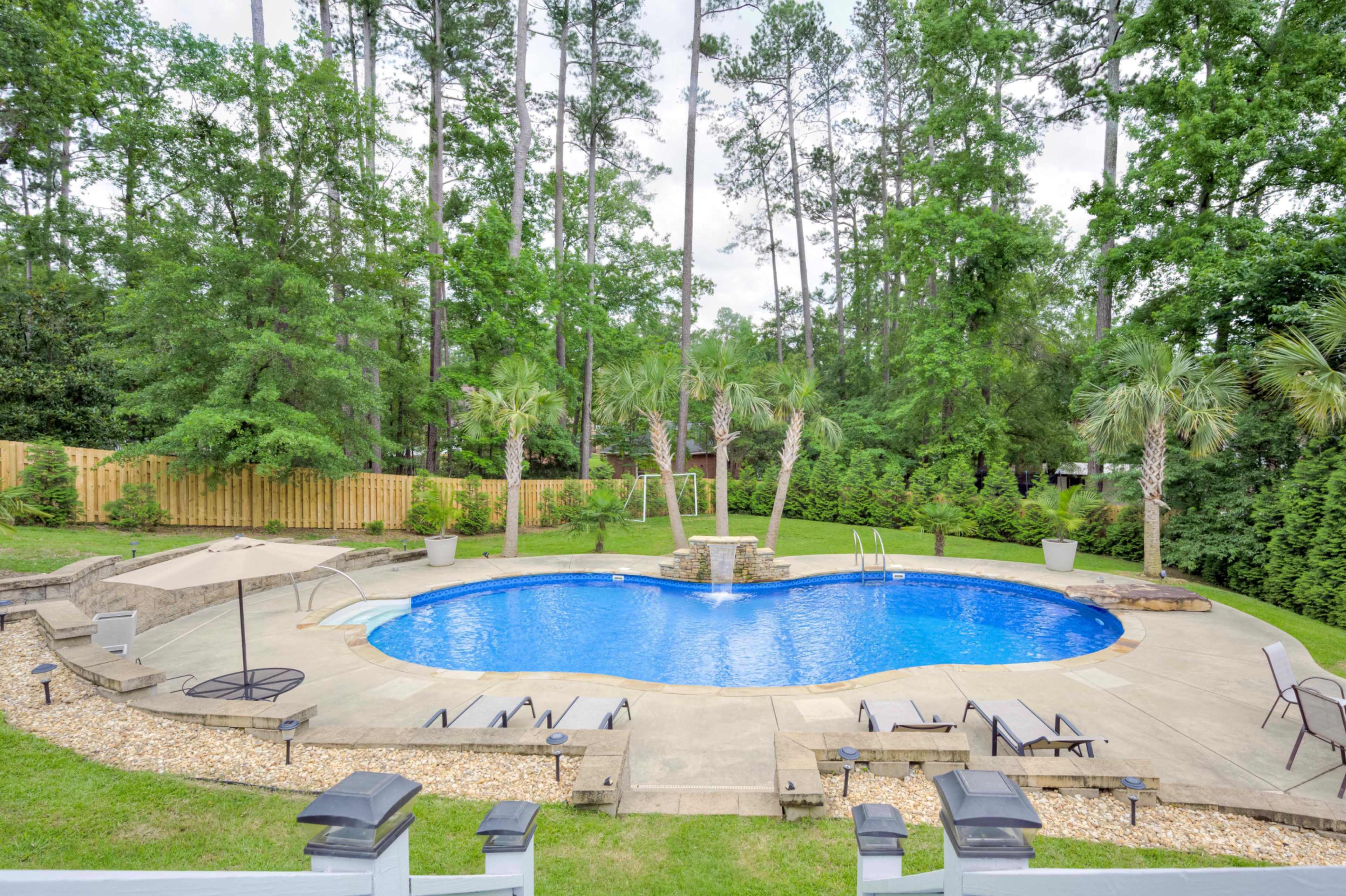 A landscaped backyard features a kidney-shaped swimming pool surrounded by stone decking, lounge chairs, and tall trees.