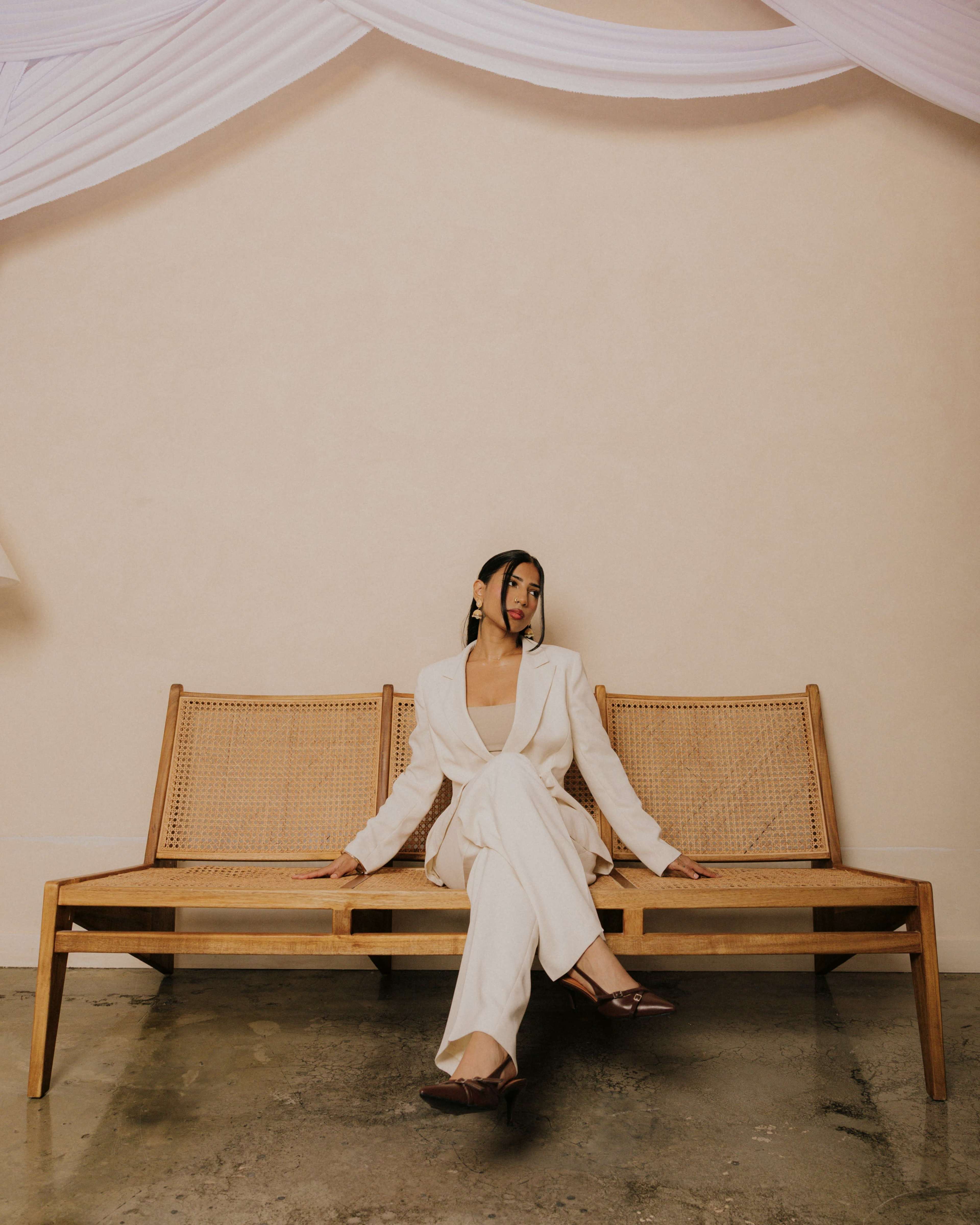 A person wearing a white suit sits on a wooden bench against a plain wall.