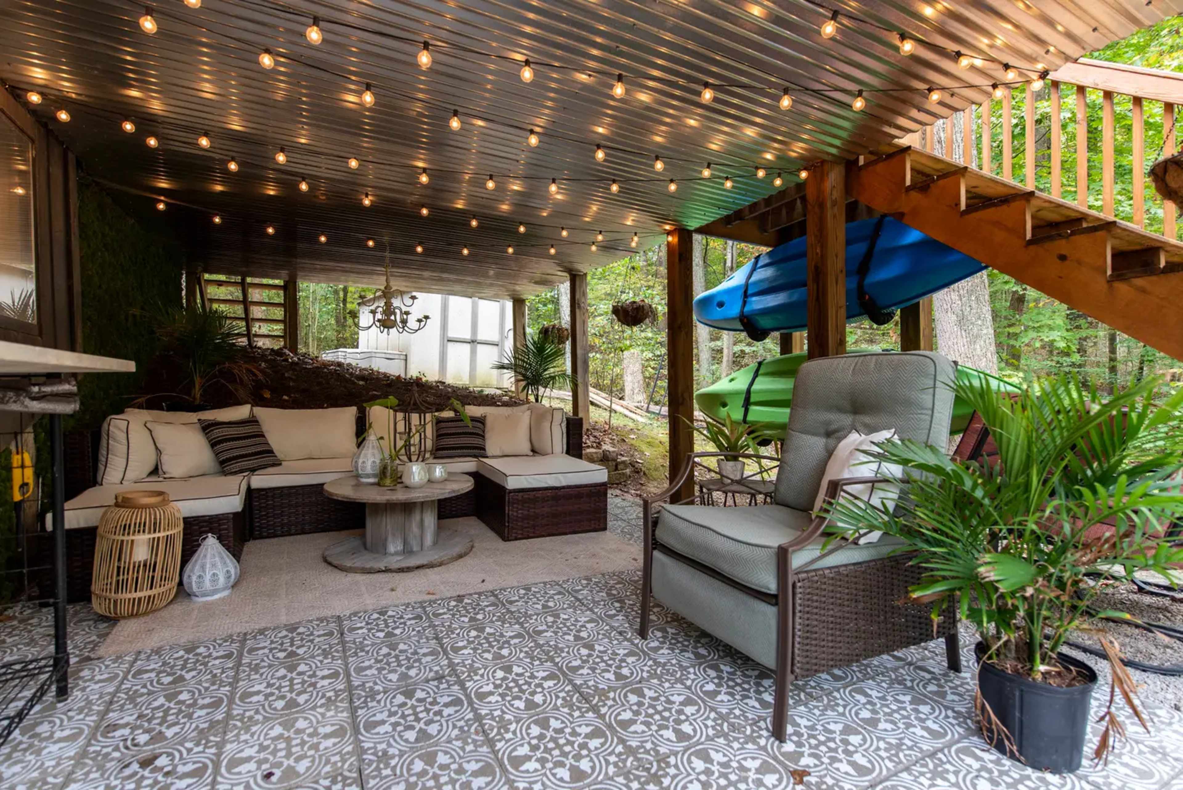 The image shows a cozy outdoor seating area under a deck, featuring a sofa set, decorative plants, and string lights overhead.