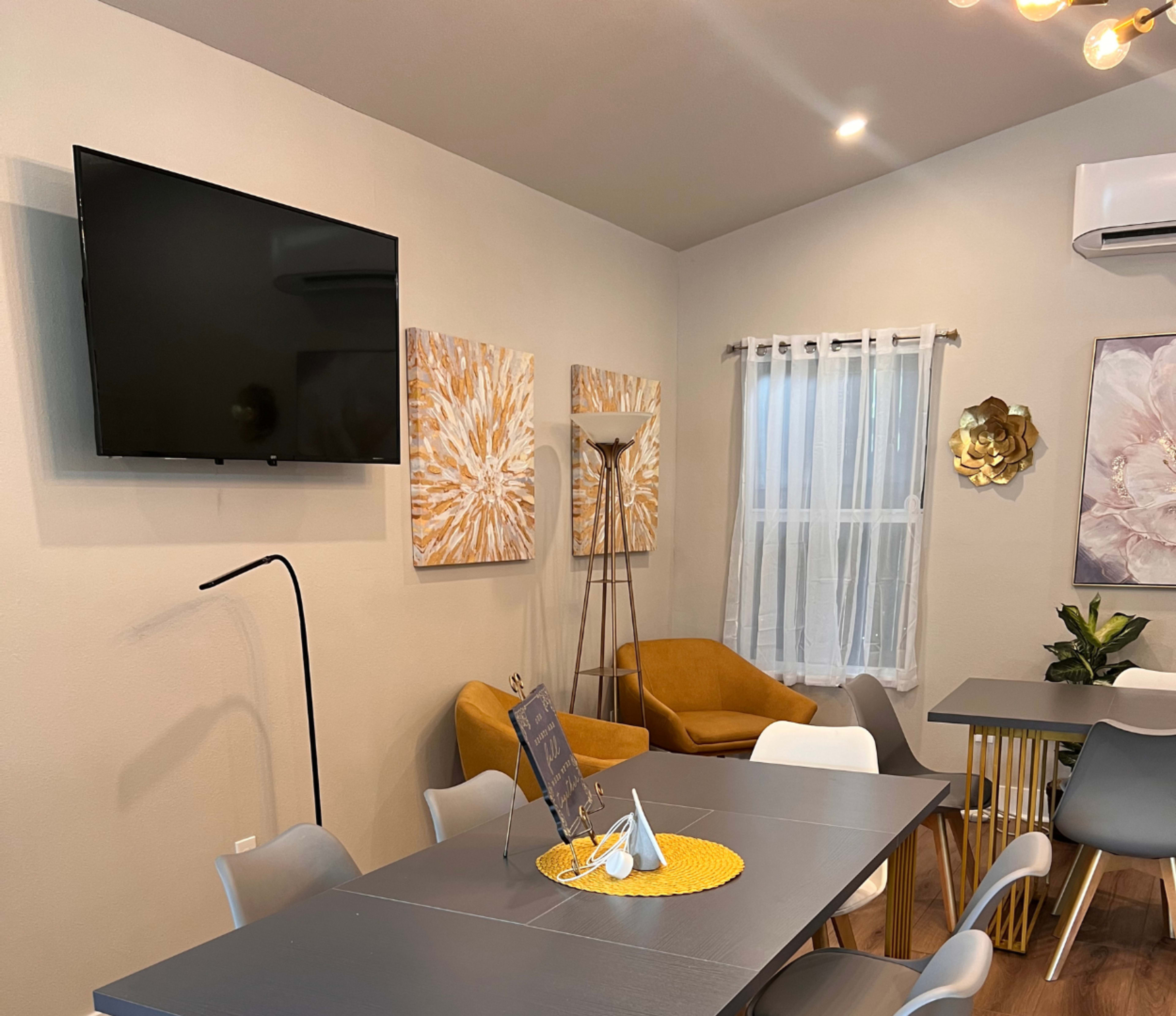 A living space features a wall-mounted television, two abstract art pieces, a mustard yellow chair, a dining table with gray chairs, and a decorative centerpiece.