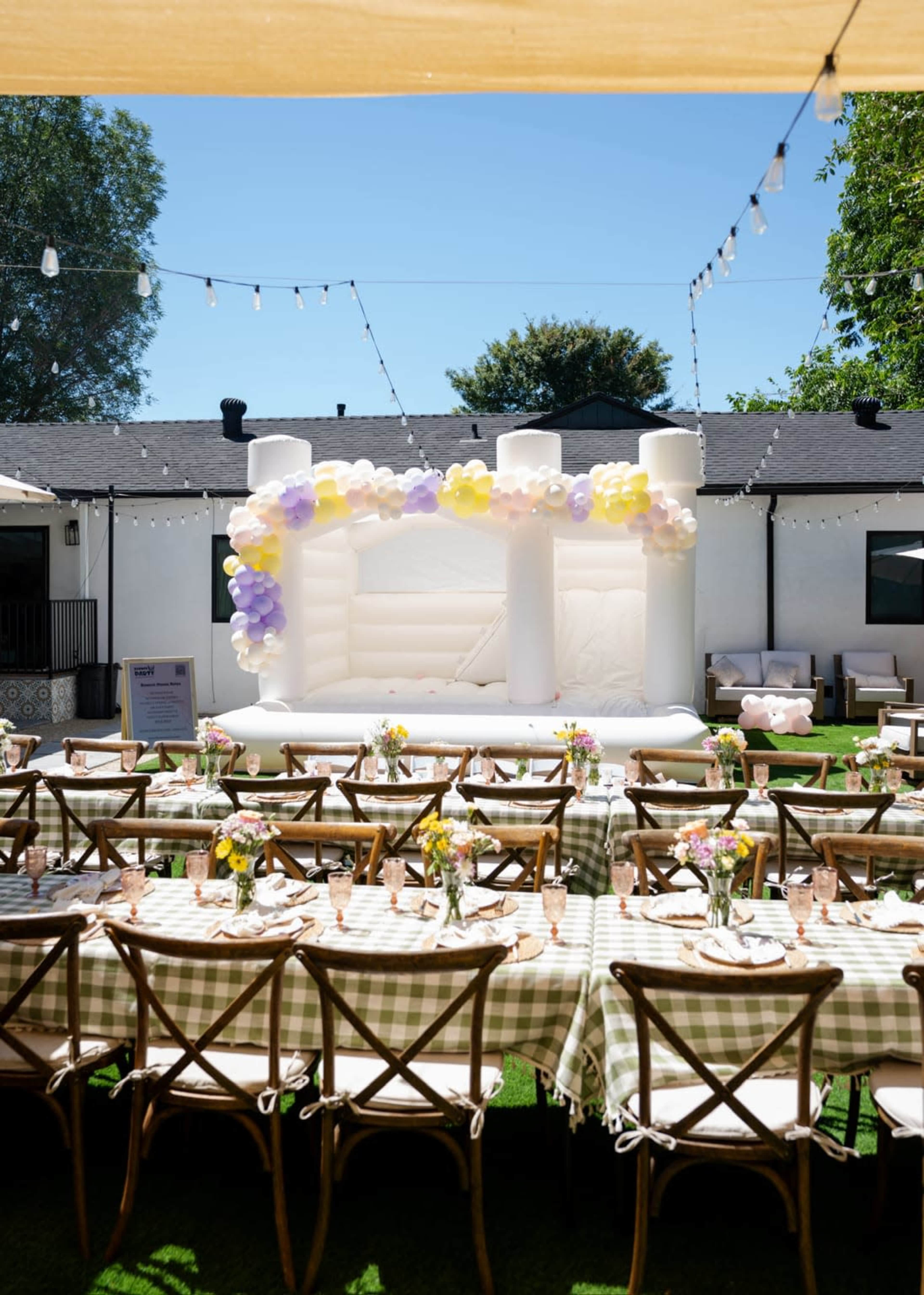 The image shows a backyard party setting with a large inflatable bounce house decorated with balloons and a long table adorned with checkered tablecloths and floral centerpieces.