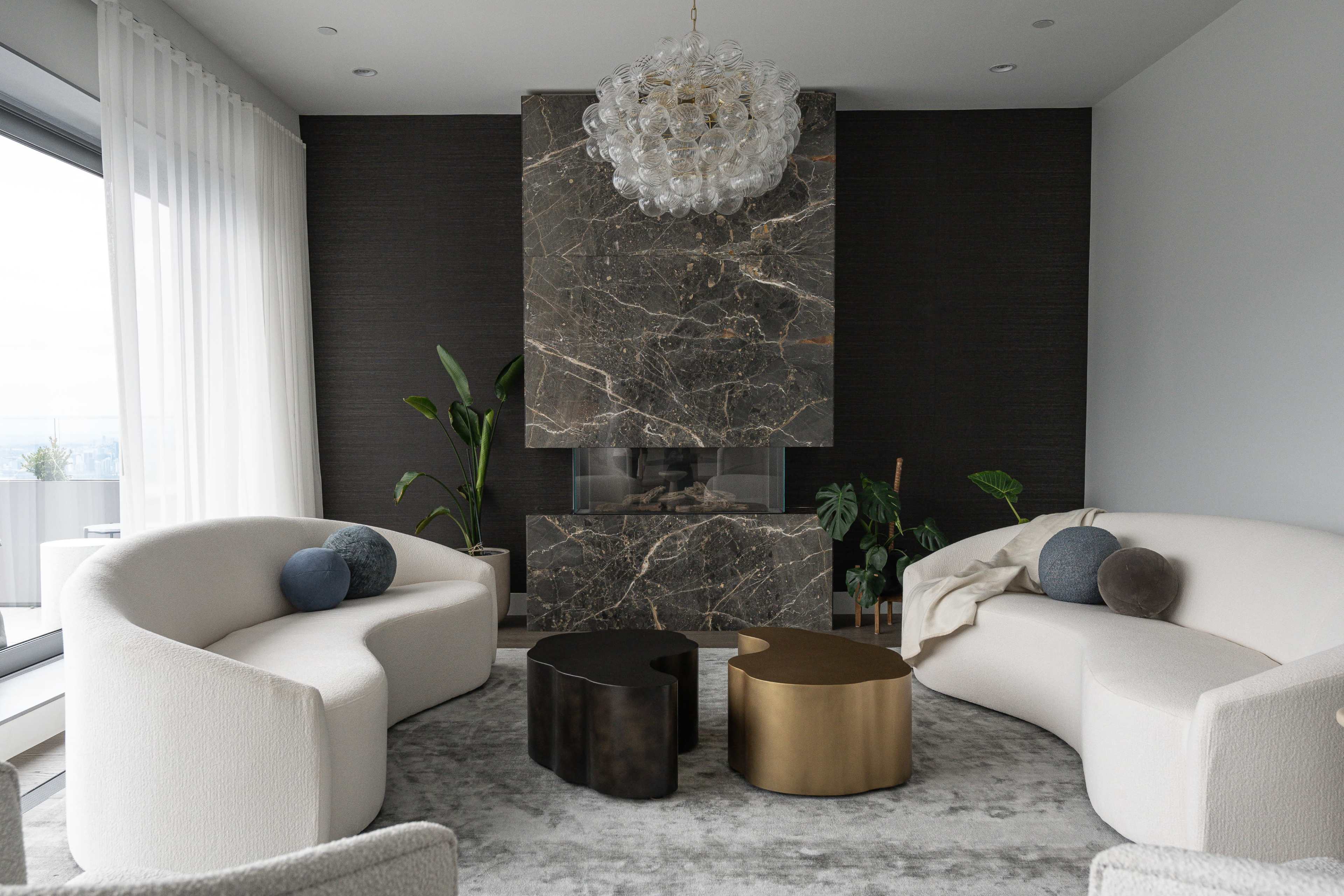 The image shows a modern living room featuring two curved sofas facing a central fireplace with a marble feature wall, accompanied by two abstract coffee tables and decorative plants.