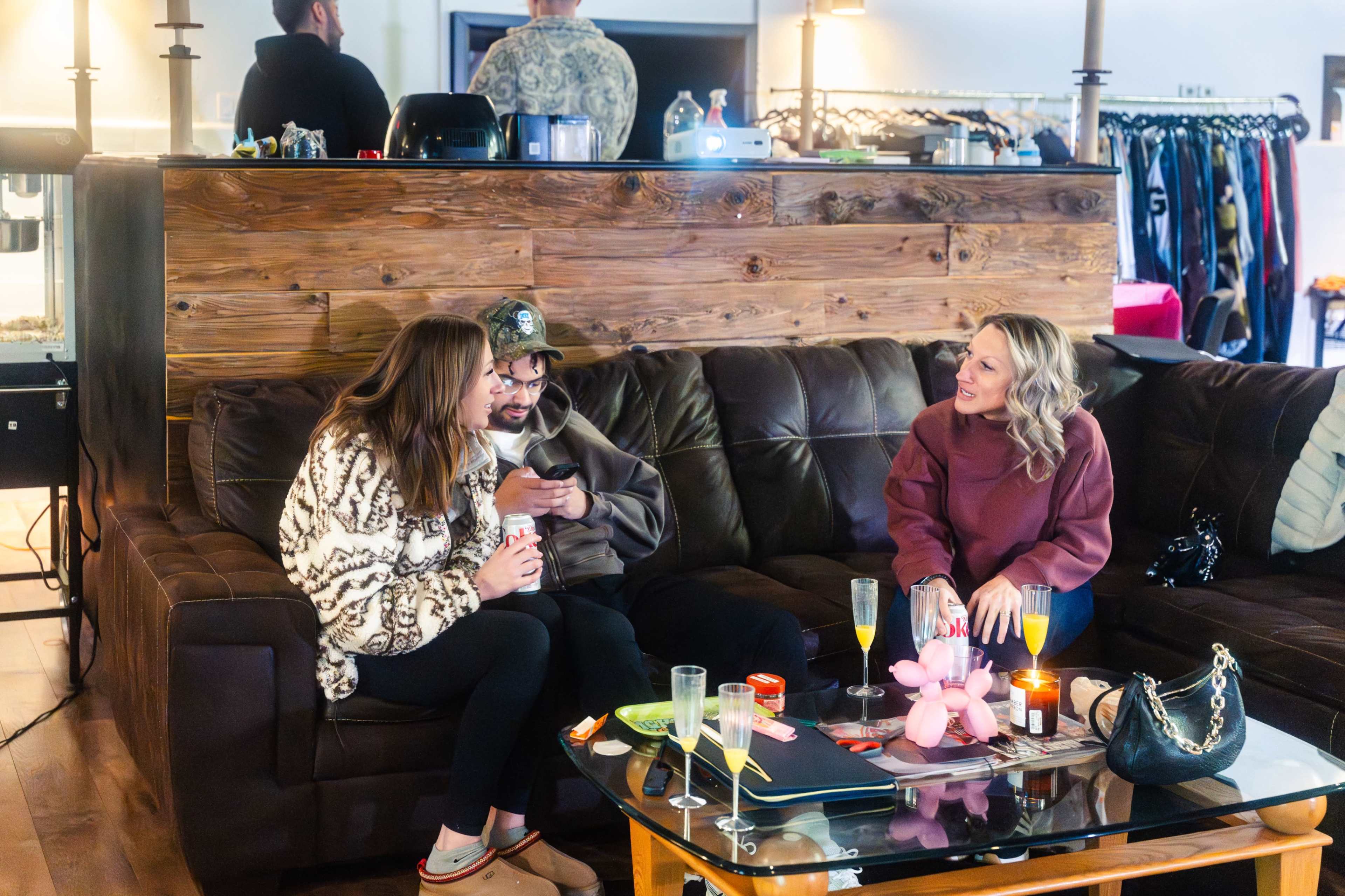 Three people sit on a brown couch in a cozy setting surrounded by clothing racks and dim lighting, engaged in conversation.