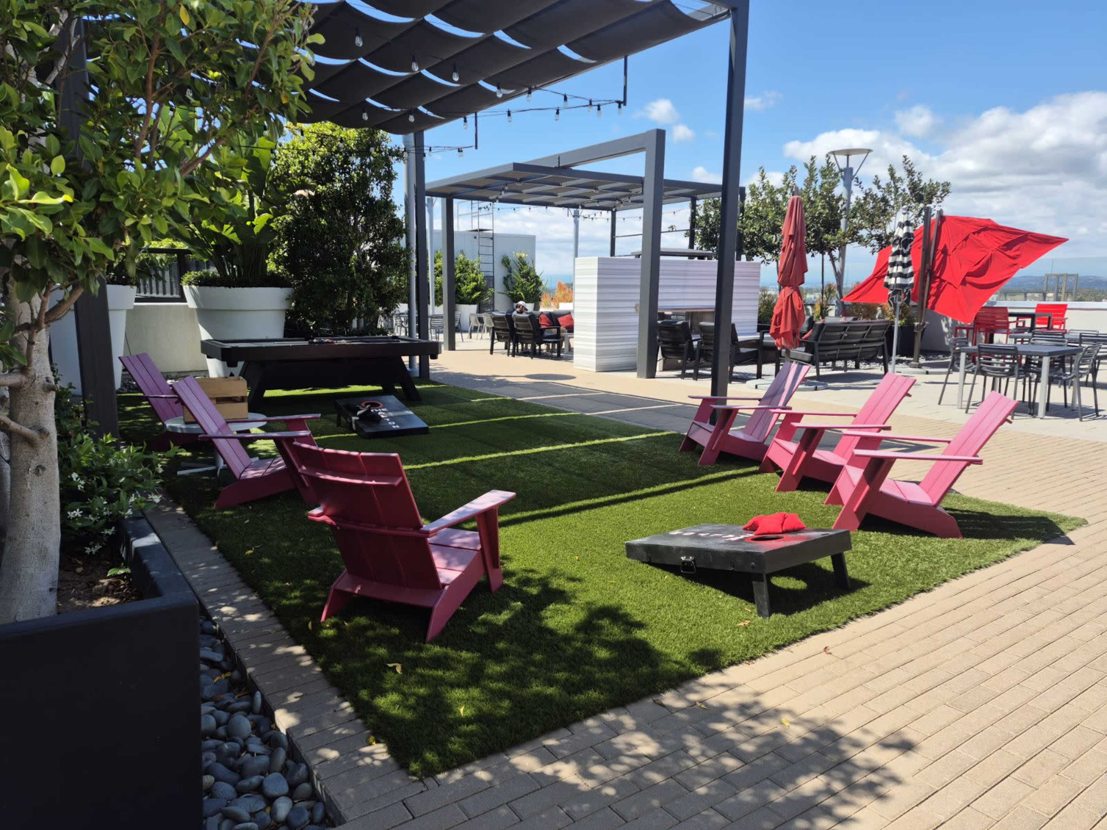 The image shows a landscaped outdoor area with pink lawn chairs arranged on artificial grass, surrounded by potted plants and patio furniture beneath a pergola.