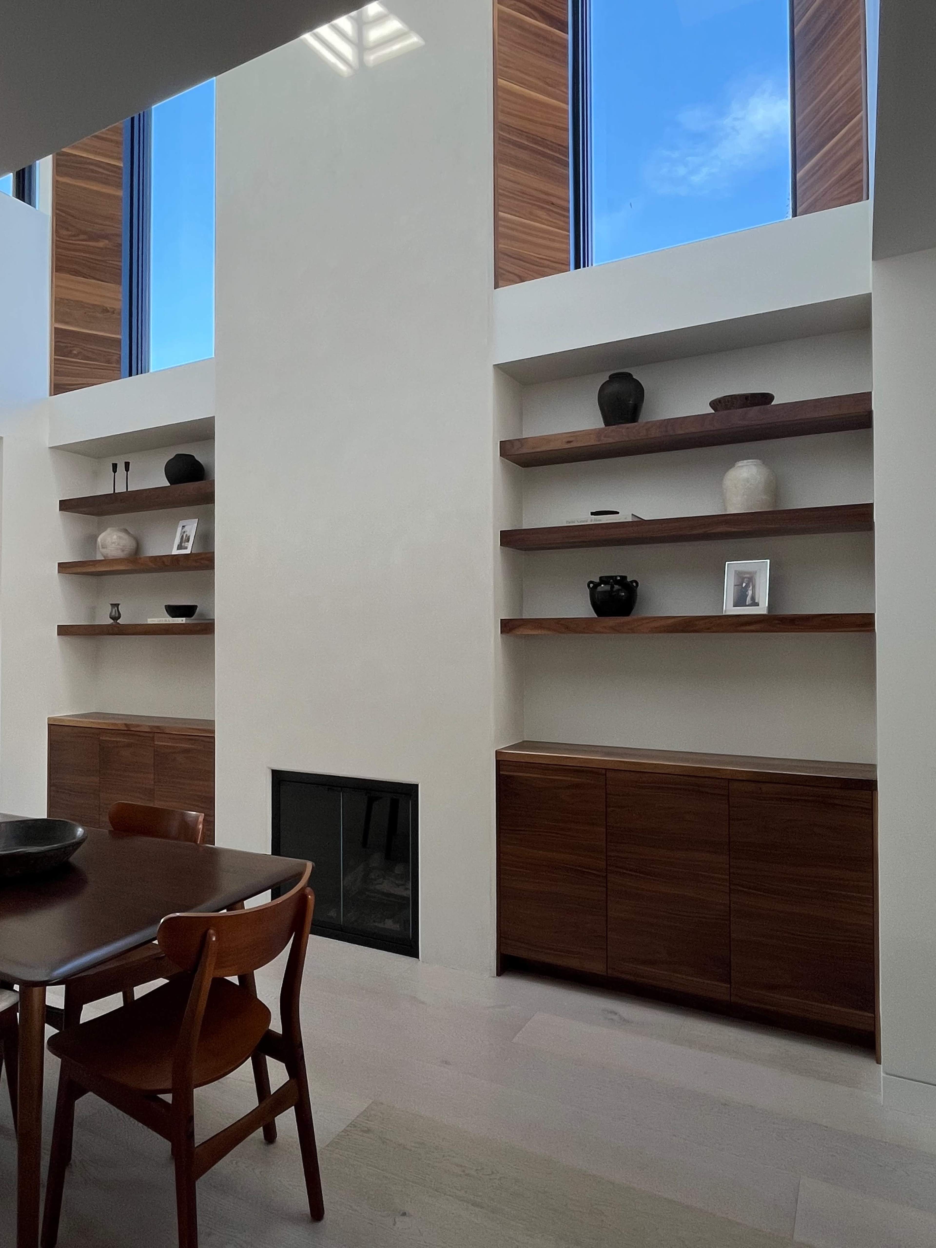The image shows a modern interior with tall windows, wooden shelves displaying decor, and a dining table with wooden chairs.