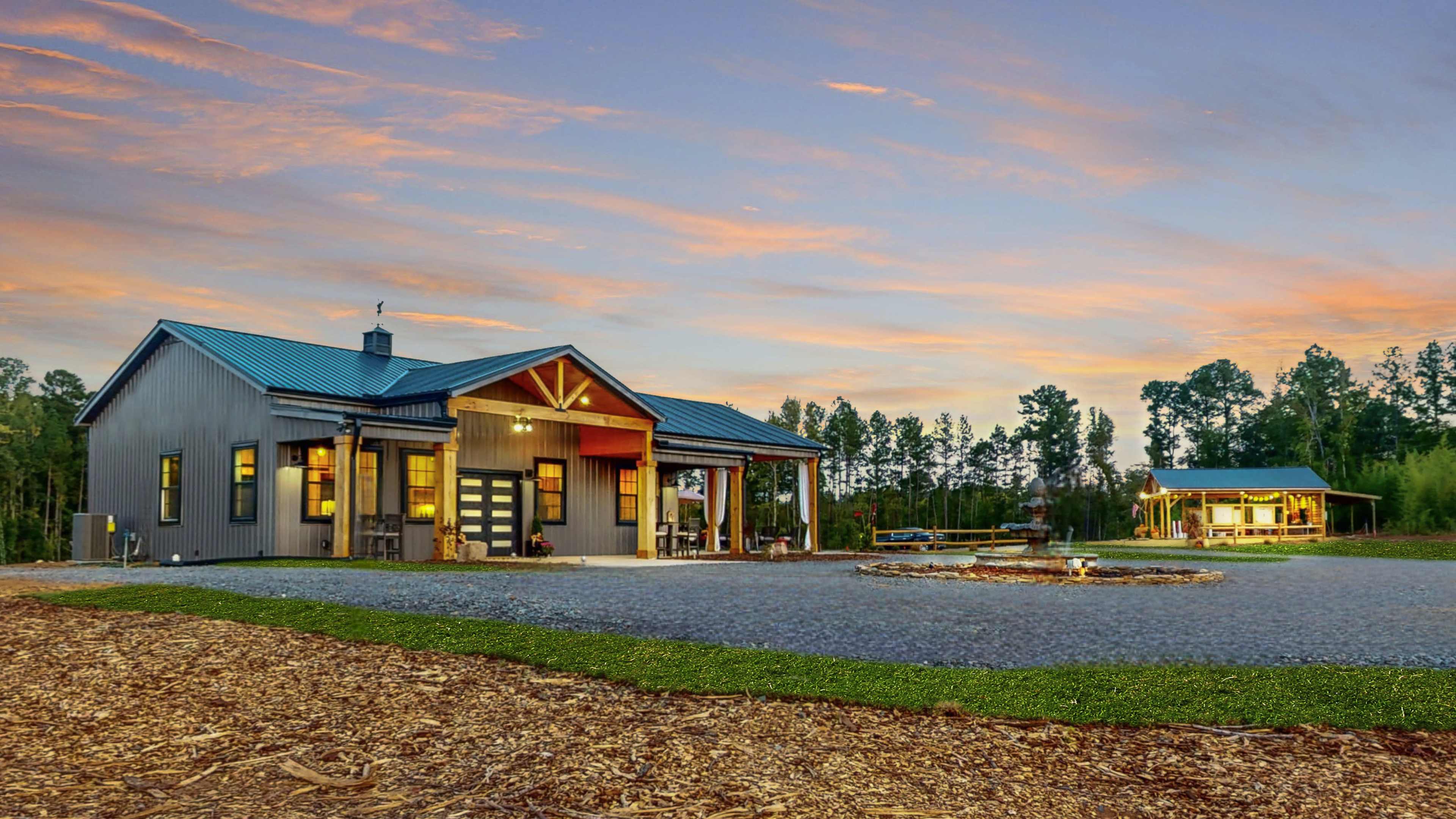 A modern house with a metal roof and wooden accents is pictured beside a circular fire pit area surrounded by trees at sunset.