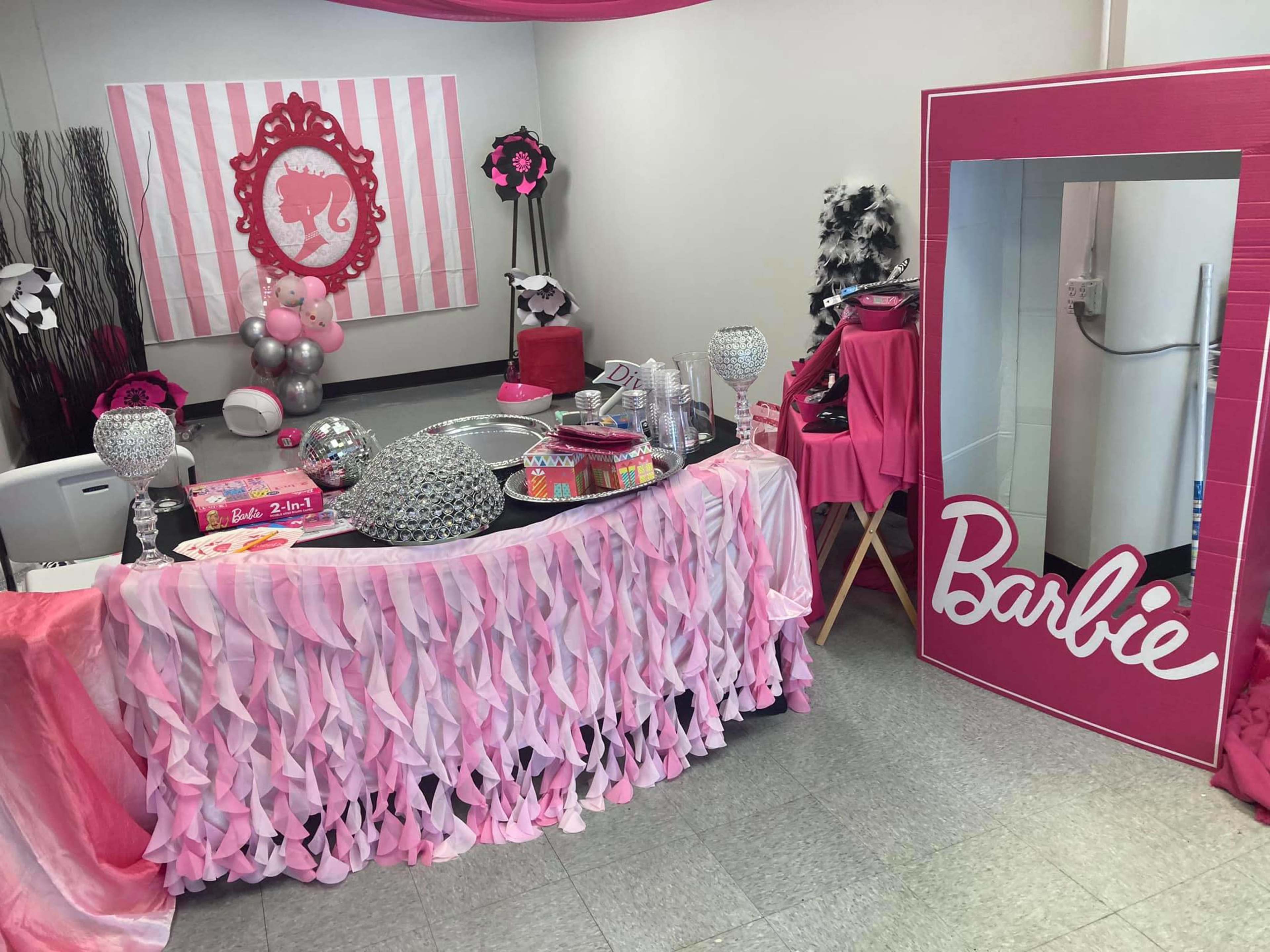 The scene features a colorful party setup decorated in pink and white, including a table with various party supplies, a large Barbie-themed mirror, and balloon decorations.