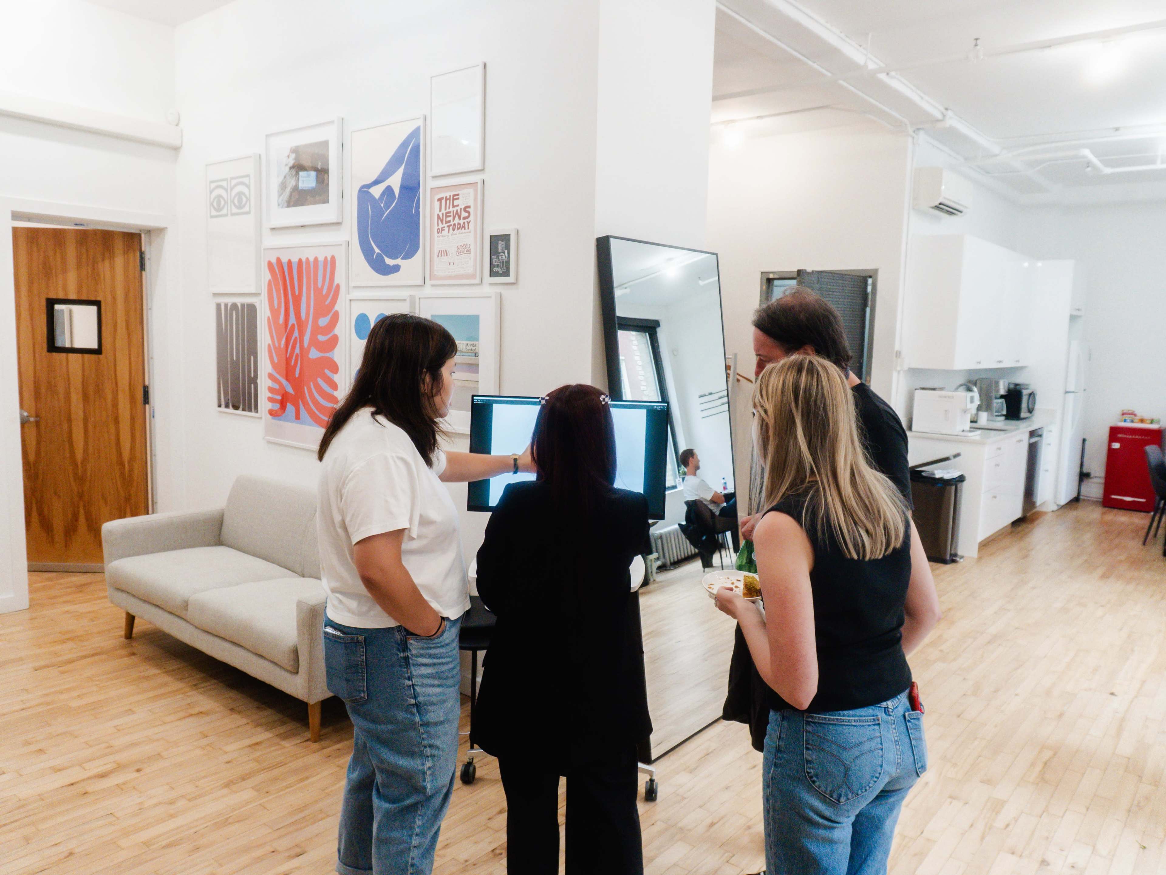 Four people gather around a computer screen in a modern office space, discussing something while a kitchen area and sofa are visible in the background.