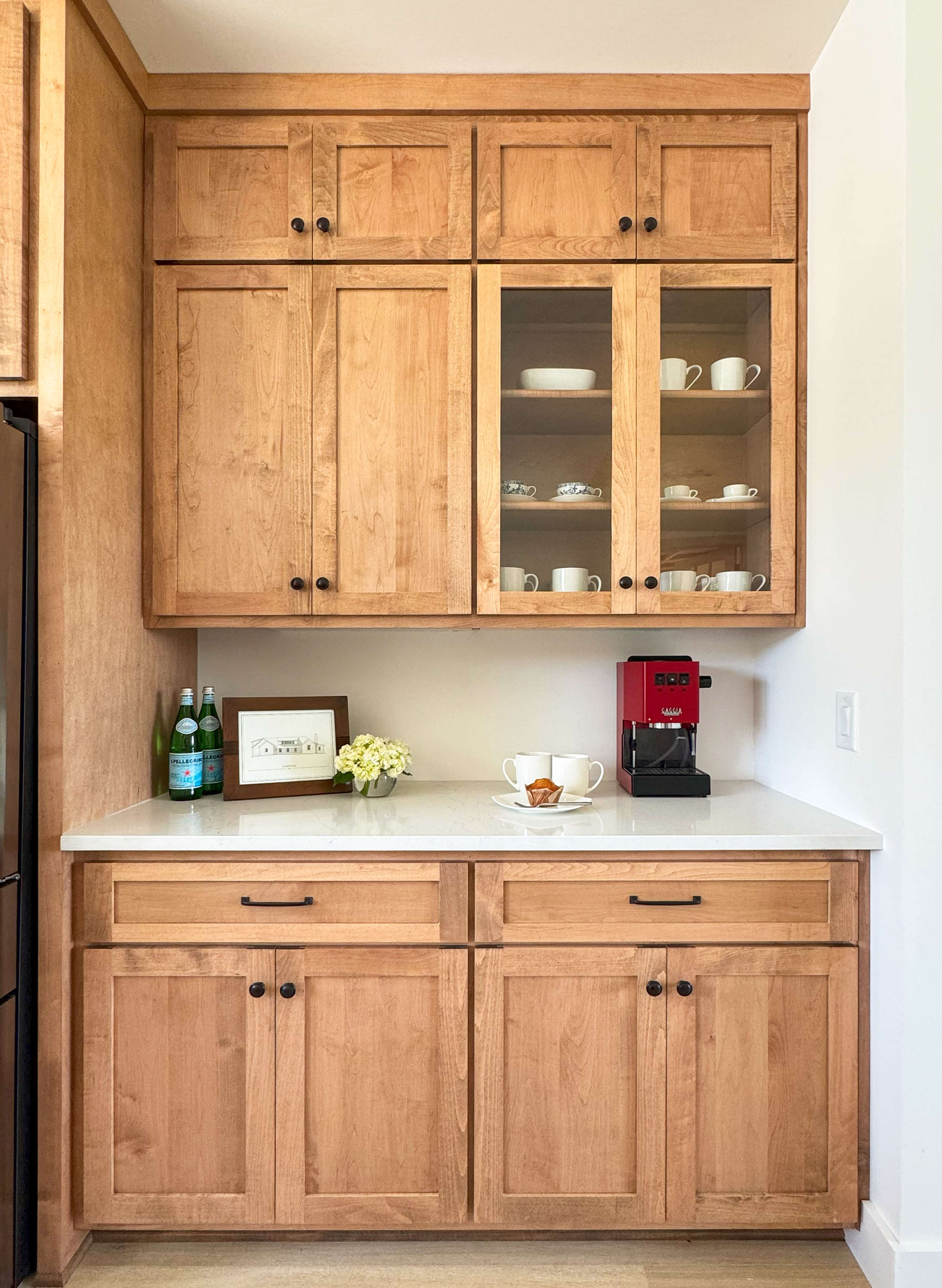 The image shows a wooden kitchen cabinet with glass-front doors, containing dishware, alongside a countertop with a coffee machine, a framed picture, and a small vase of flowers.