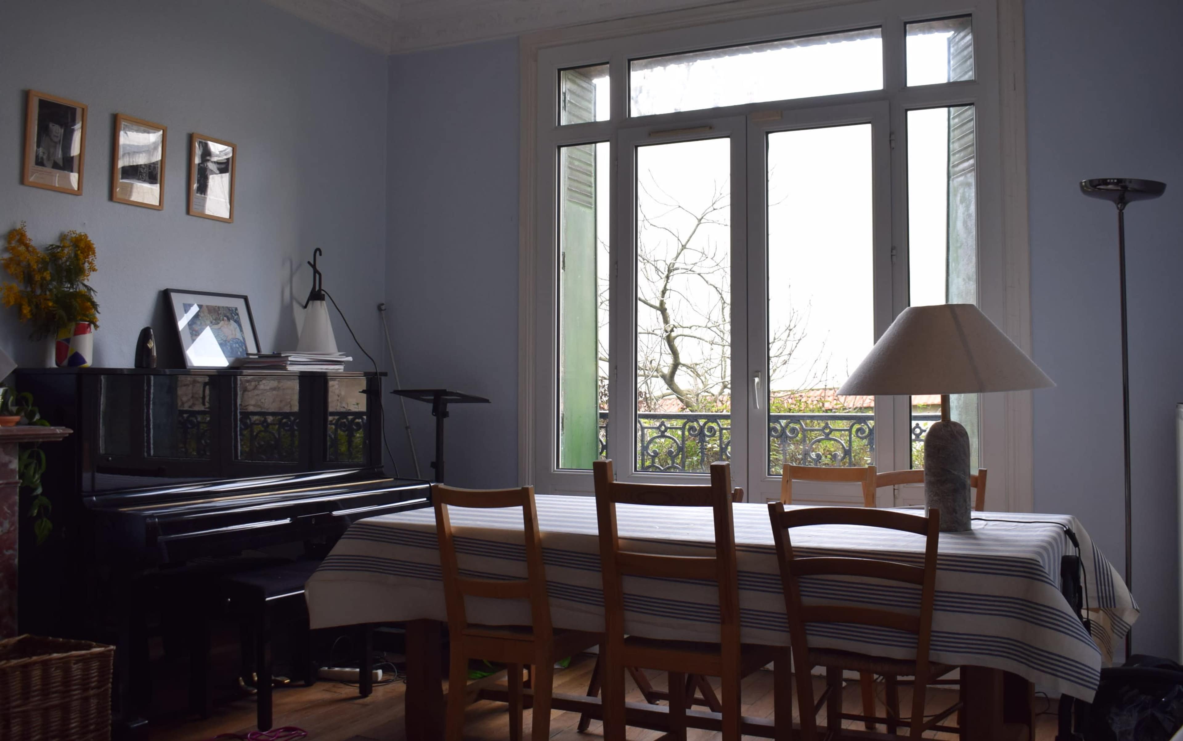The image shows a dining area with a wooden table surrounded by chairs, a piano in the corner, and large windows offering a view of a tree-lined exterior.