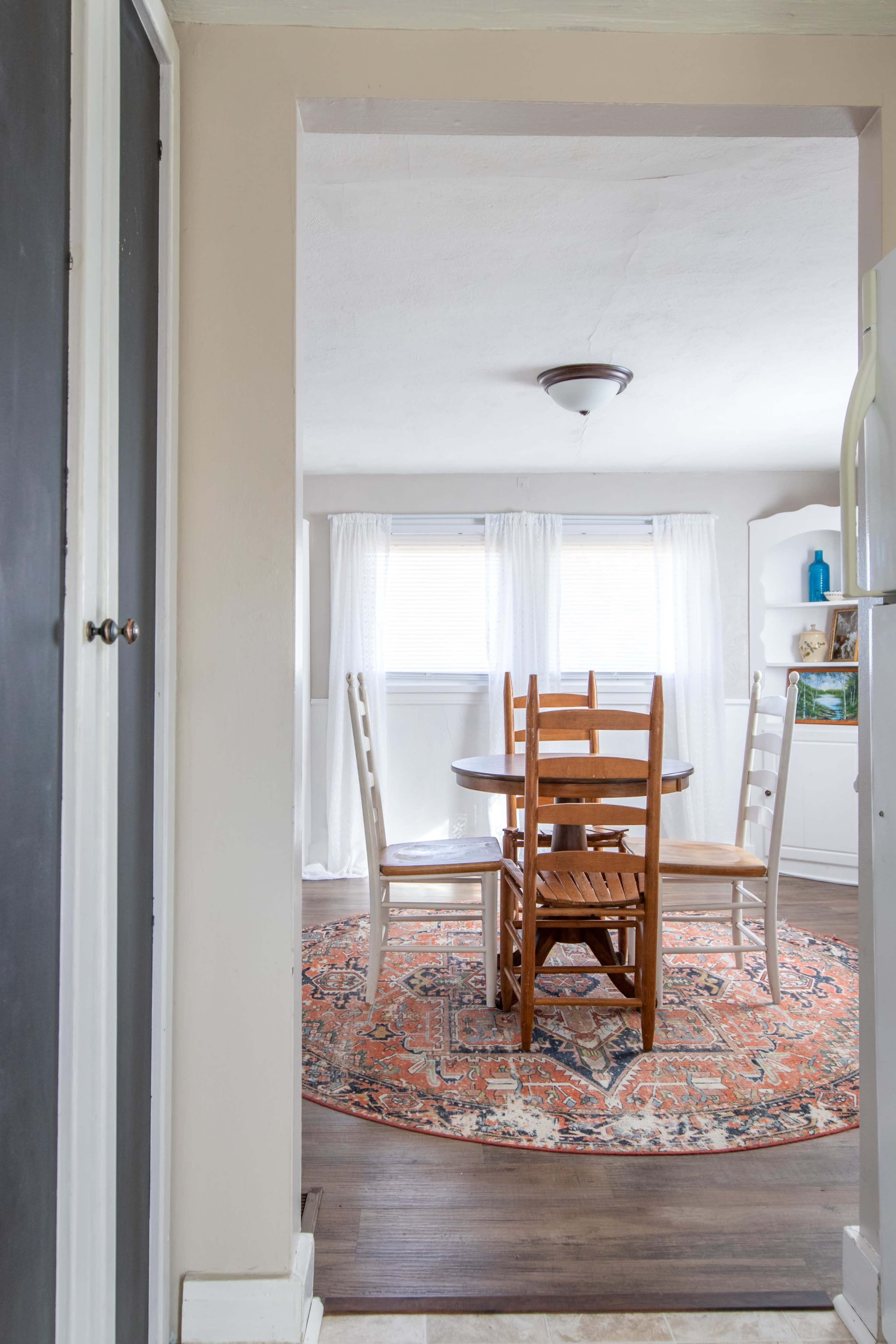A dining area is visible through an open doorway, featuring a round wooden table surrounded by chairs, with a patterned rug beneath and natural light coming from nearby windows.