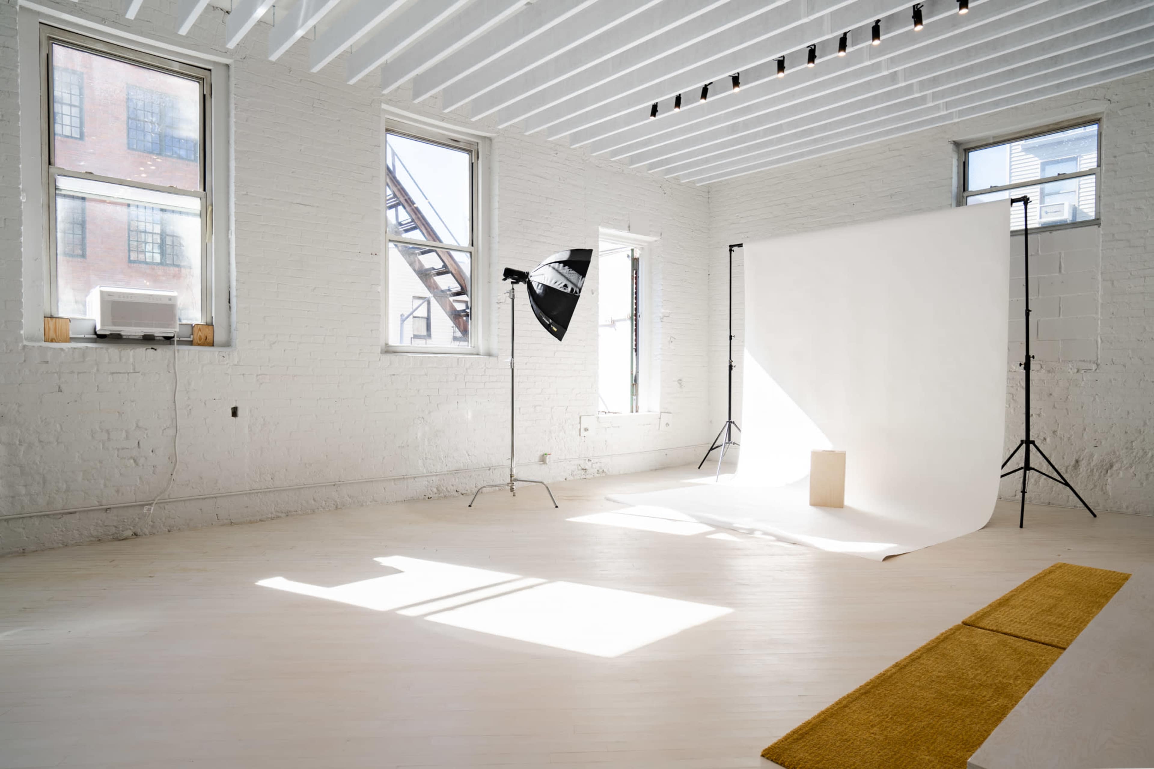 The image shows a well-lit, empty studio space with white walls, large windows, and a backdrop set up for photography.