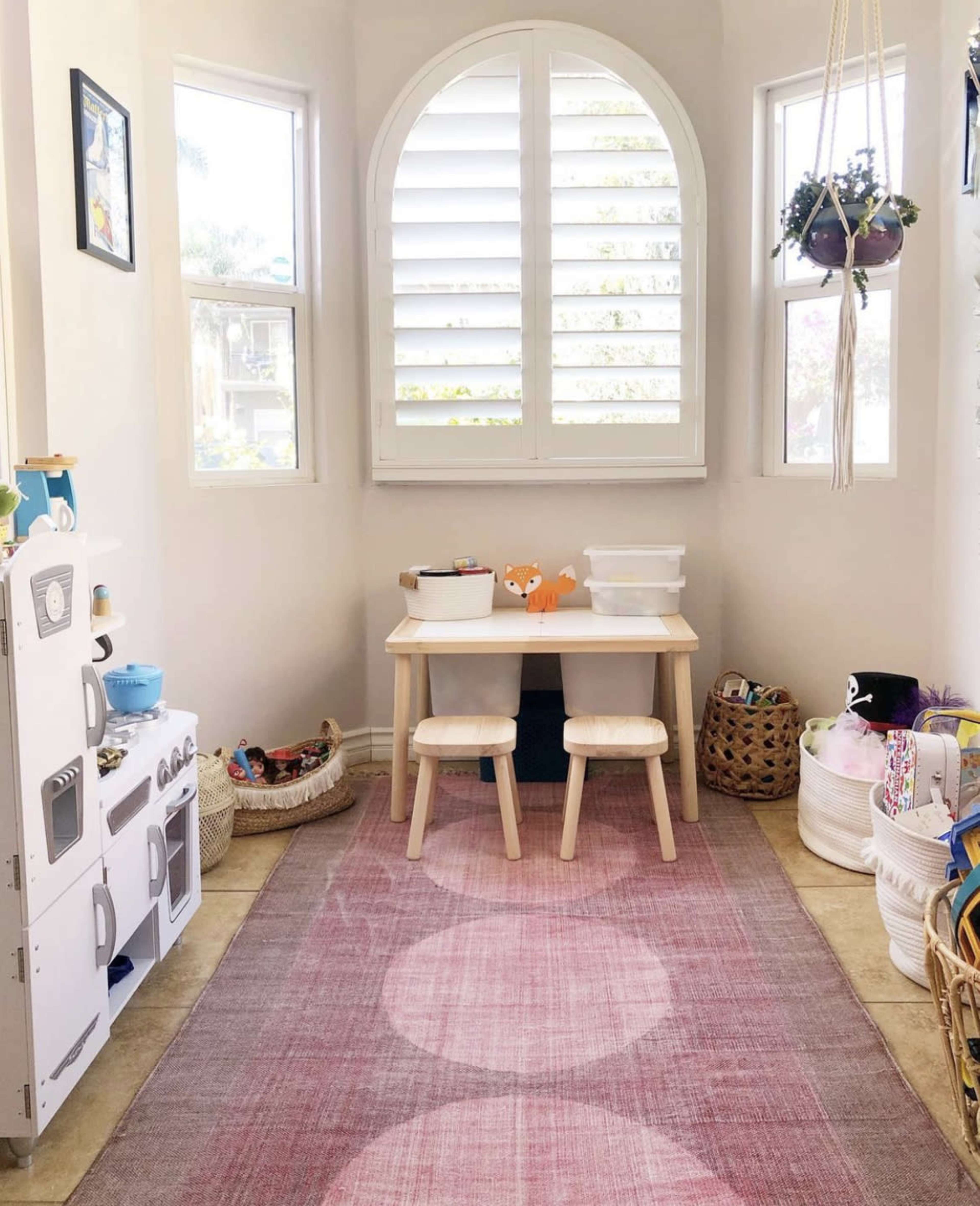 A small play area features a table with two stools, surrounded by windows and storage baskets, with a toy kitchen set against one wall.