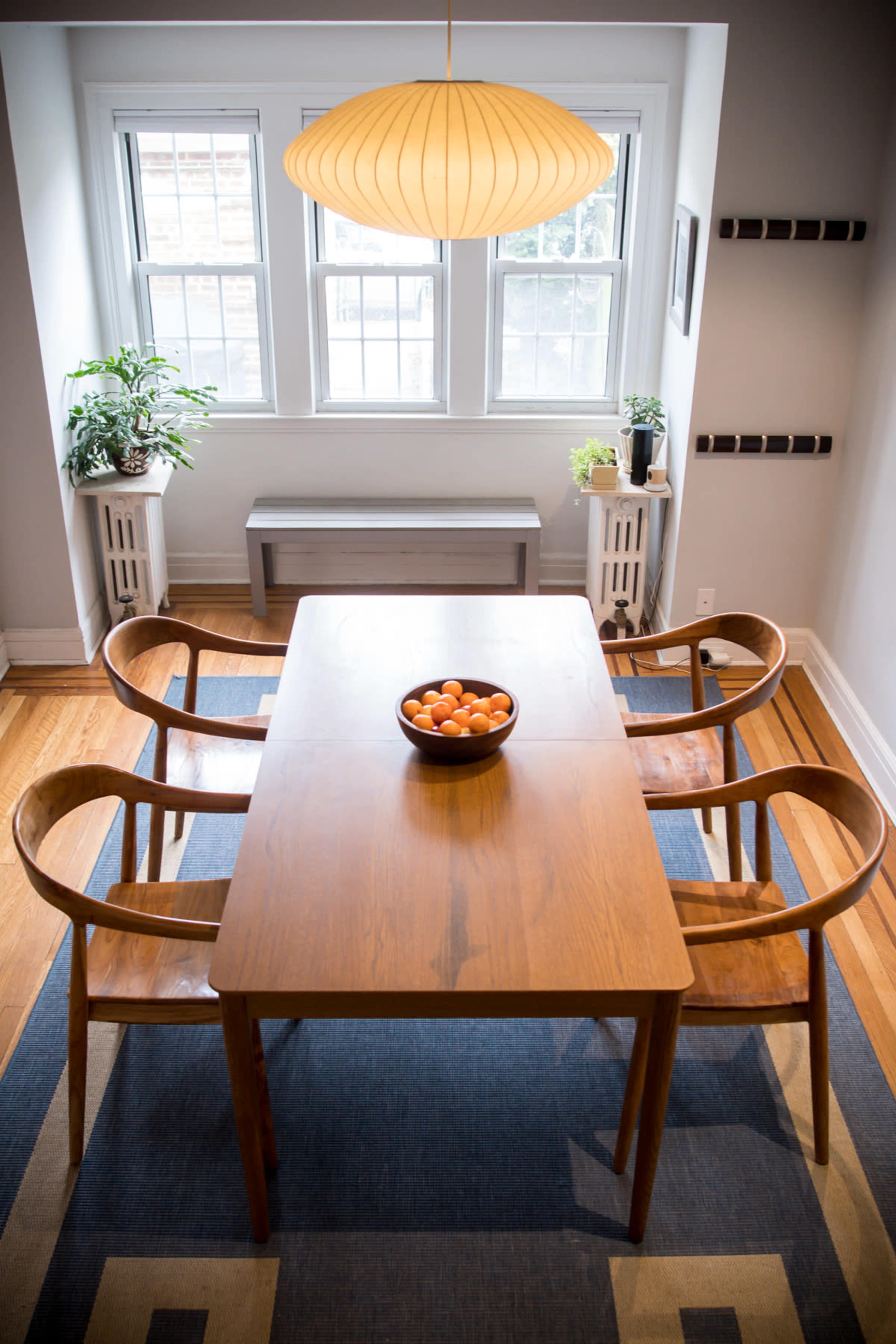 A wooden dining table with four chairs is set in a brightly lit room, featuring a bowl of oranges at its center and window plants nearby.