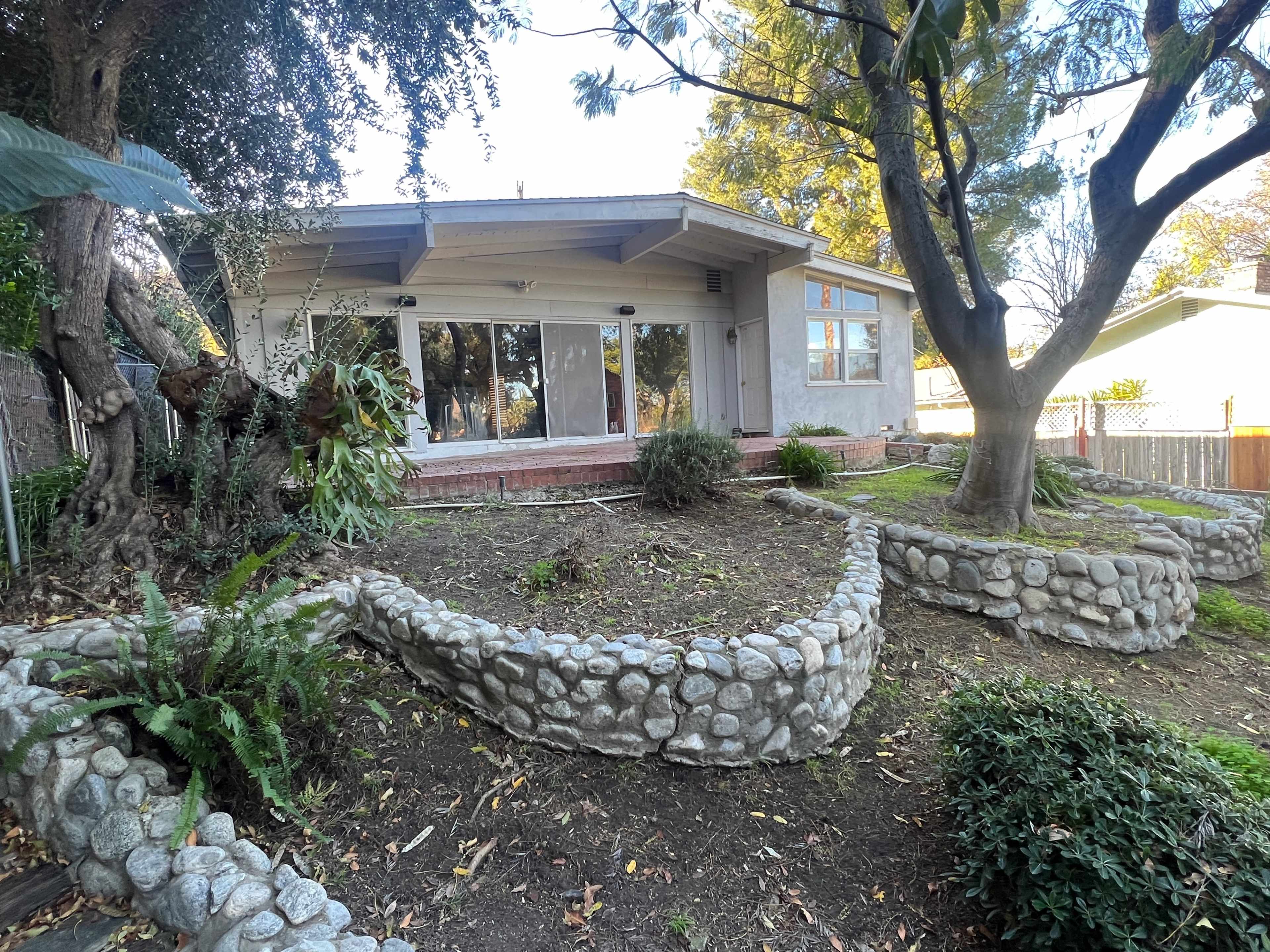 A house with large windows and a stone-bordered garden is surrounded by trees and shrubs.