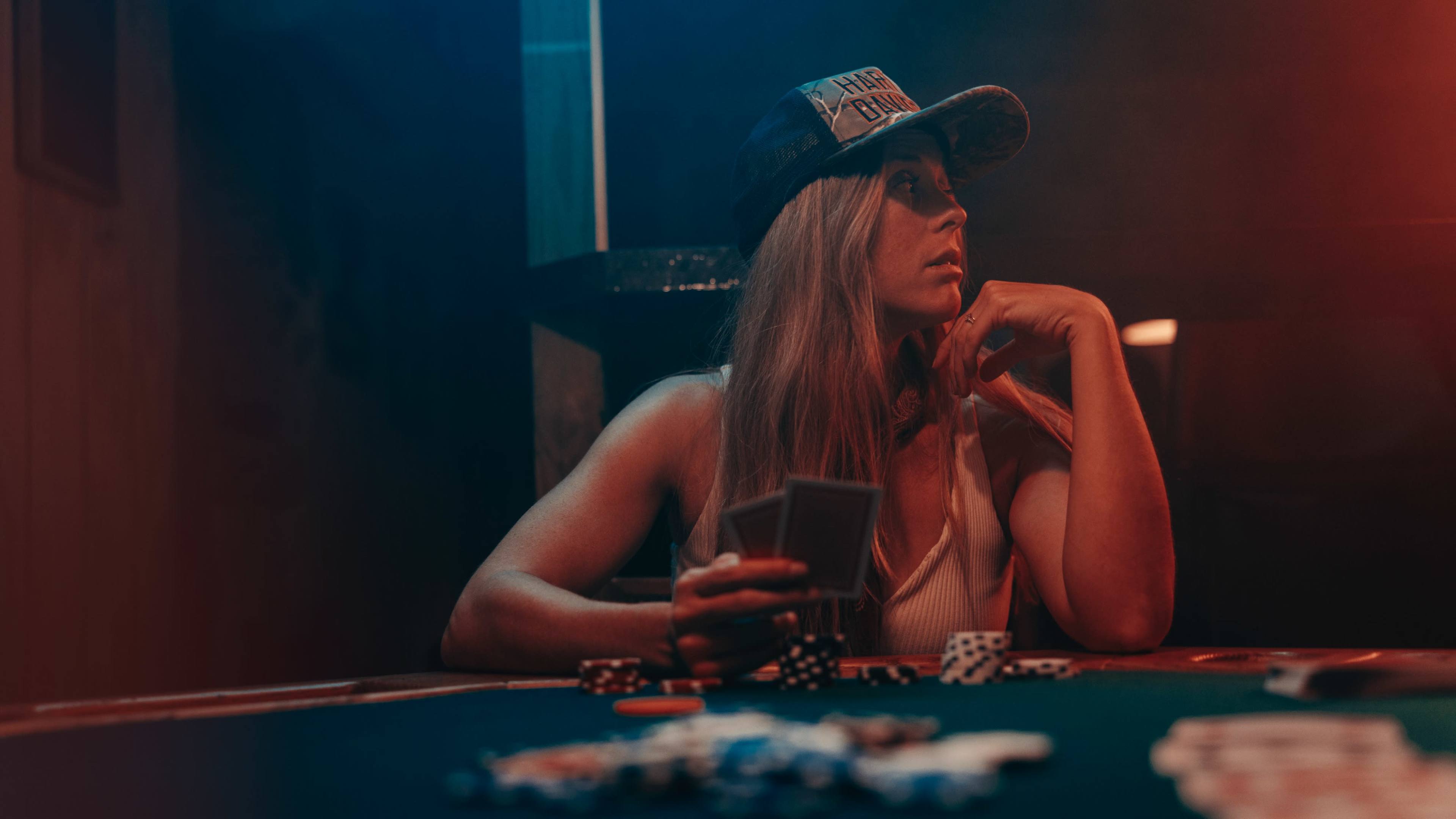 A woman holding playing cards sits at a poker table, surrounded by poker chips in a dimly lit environment.