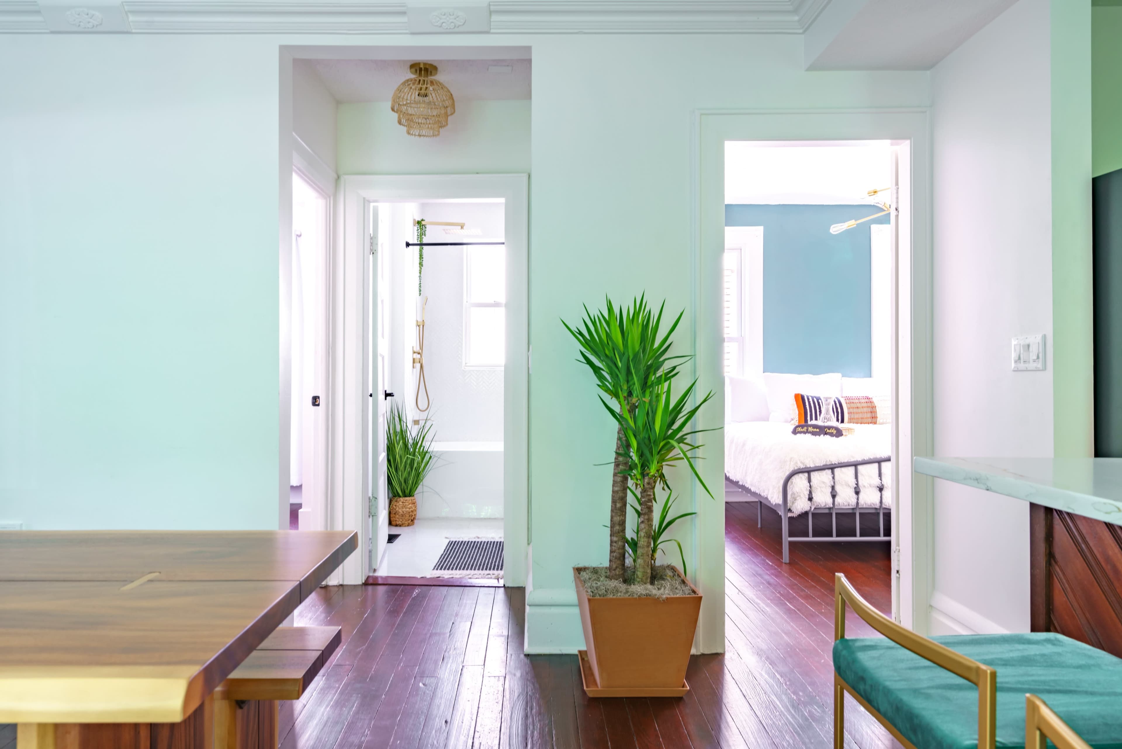 A view of a modern interior featuring a dining area with a wooden table, potted plants, and doorways leading to a bathroom and a bedroom.