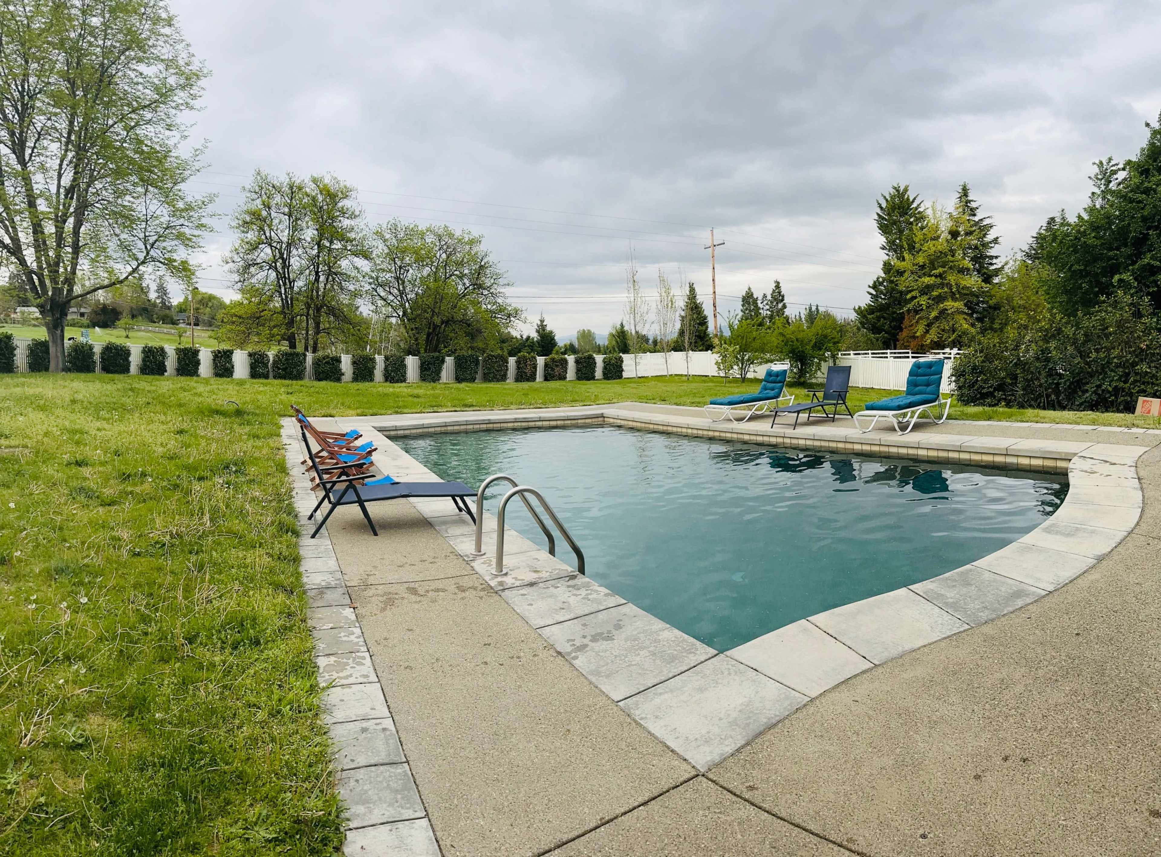 A rectangular swimming pool surrounded by a grassy area and several lounge chairs under a cloudy sky.
