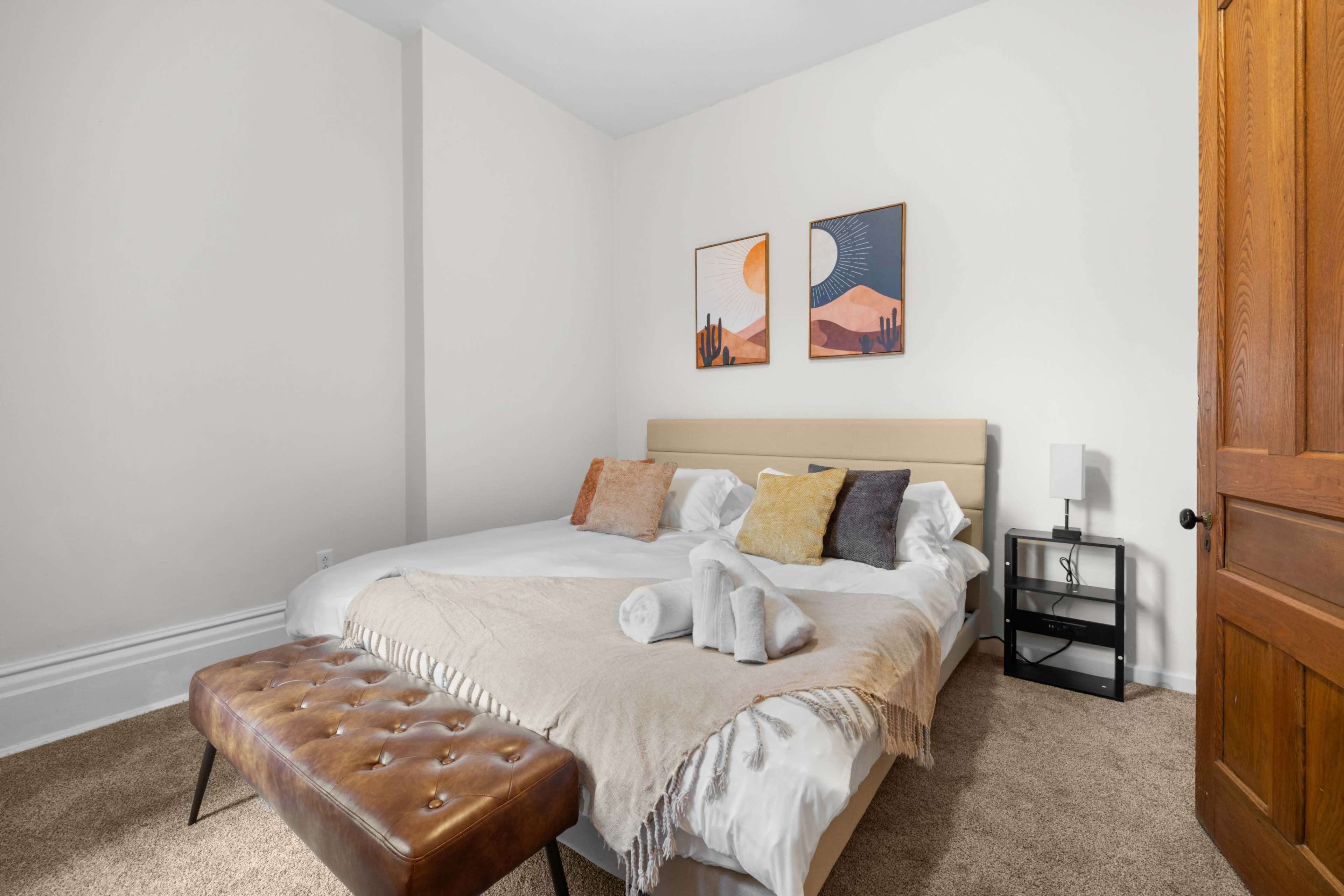 The image shows a tidy bedroom with two beds, decorative throw pillows, framed artwork on the walls, and a small bedside table with a lamp.
