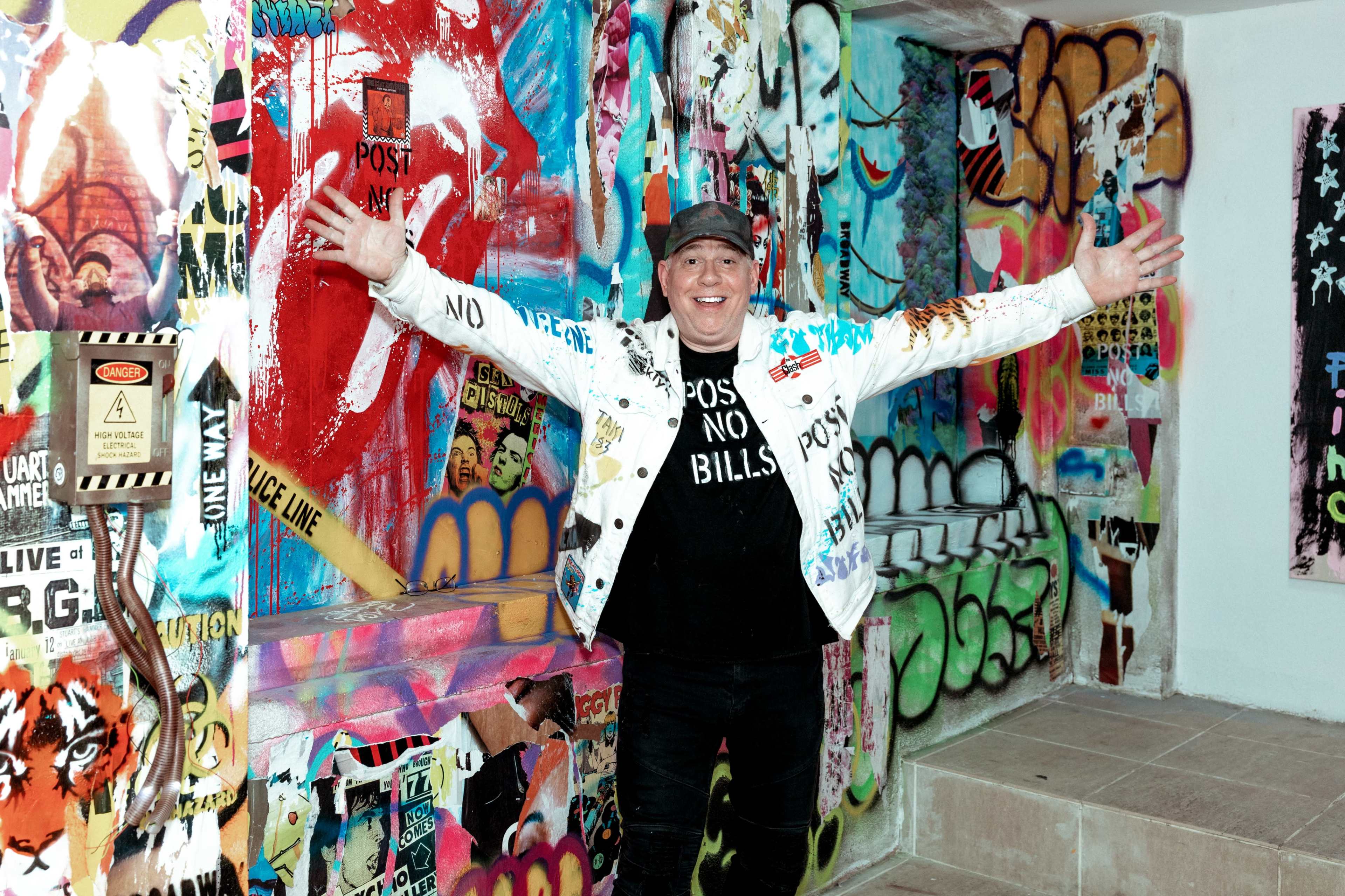 A person stands against a colorful, graffiti-covered wall with arms extended wide in a vibrant urban setting.
