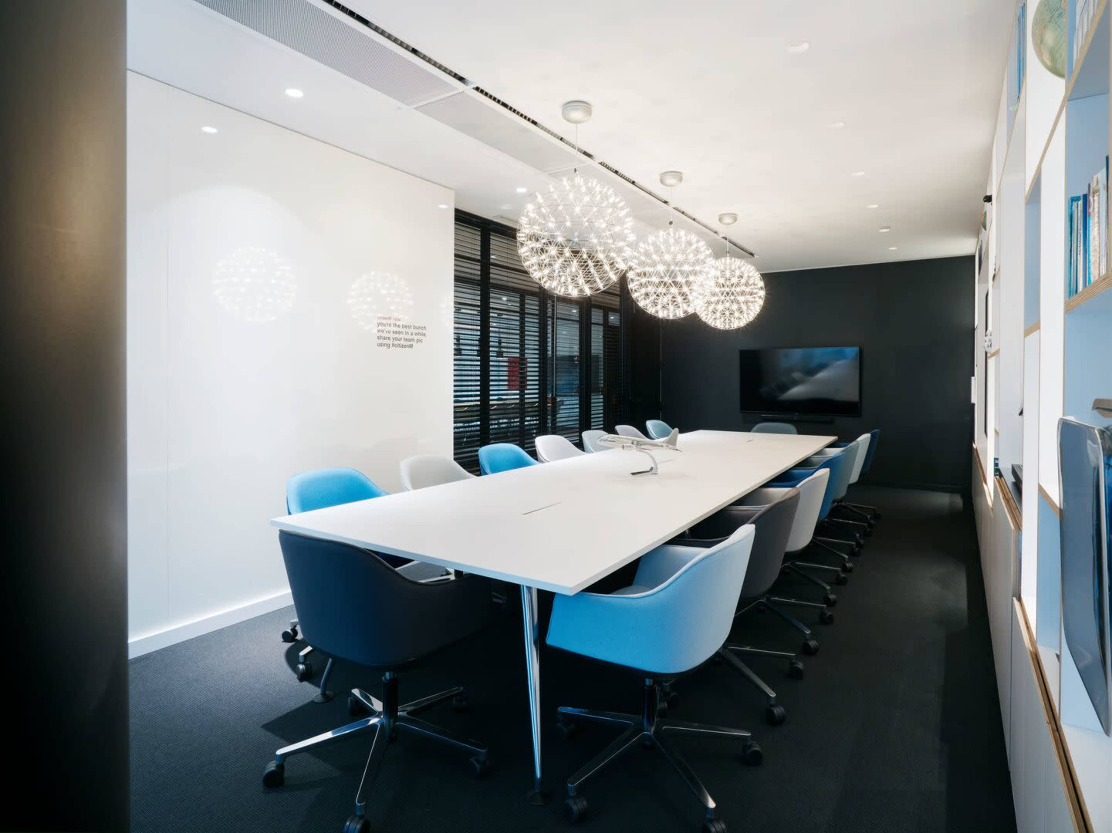 A modern conference room features a long white table surrounded by blue and gray office chairs, with large spherical light fixtures overhead and a wall-mounted screen at one end.