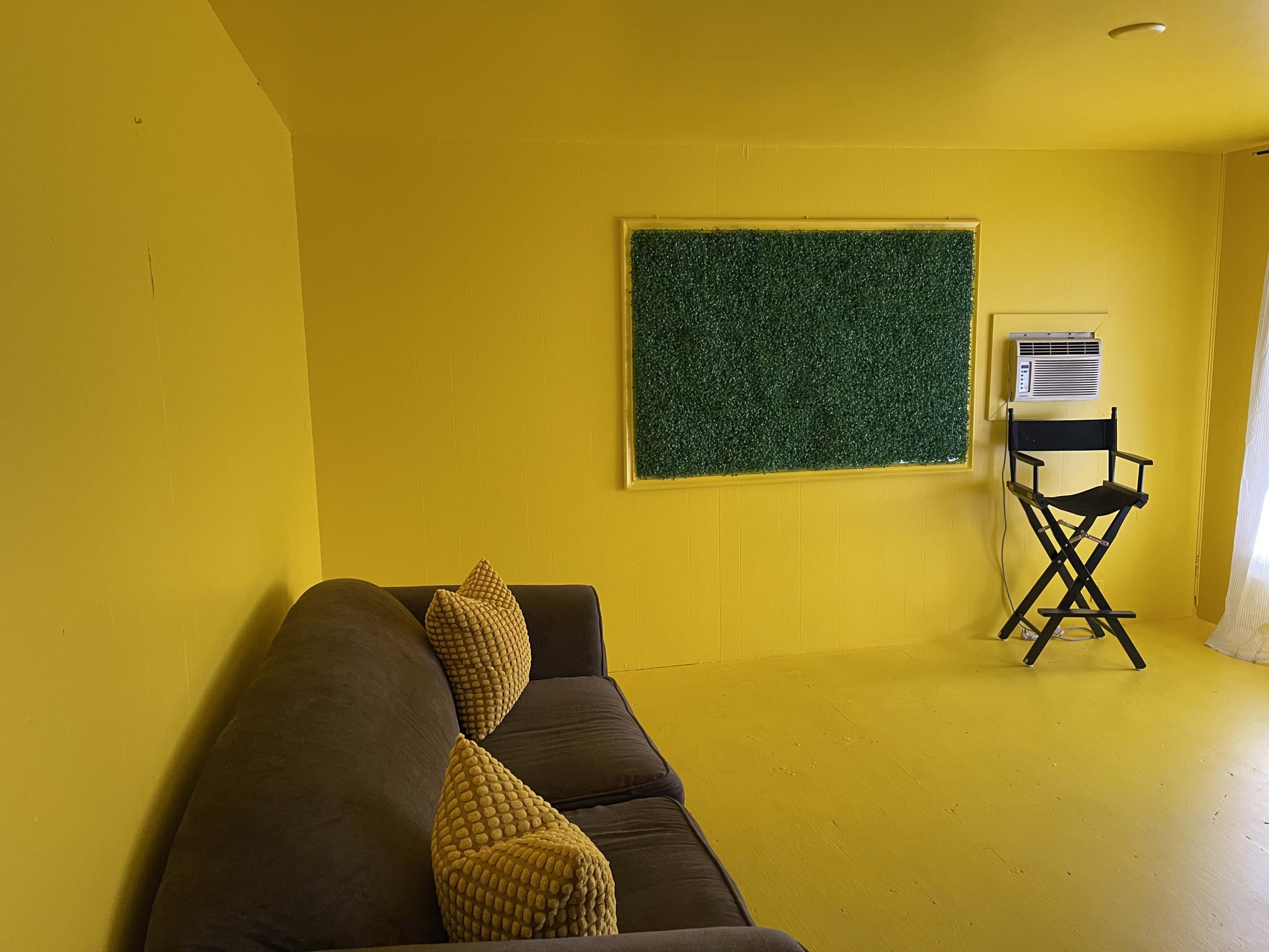 The room features yellow walls and flooring, a gray couch with yellow pillows, a green wall panel, and a director's chair next to an air conditioning unit.