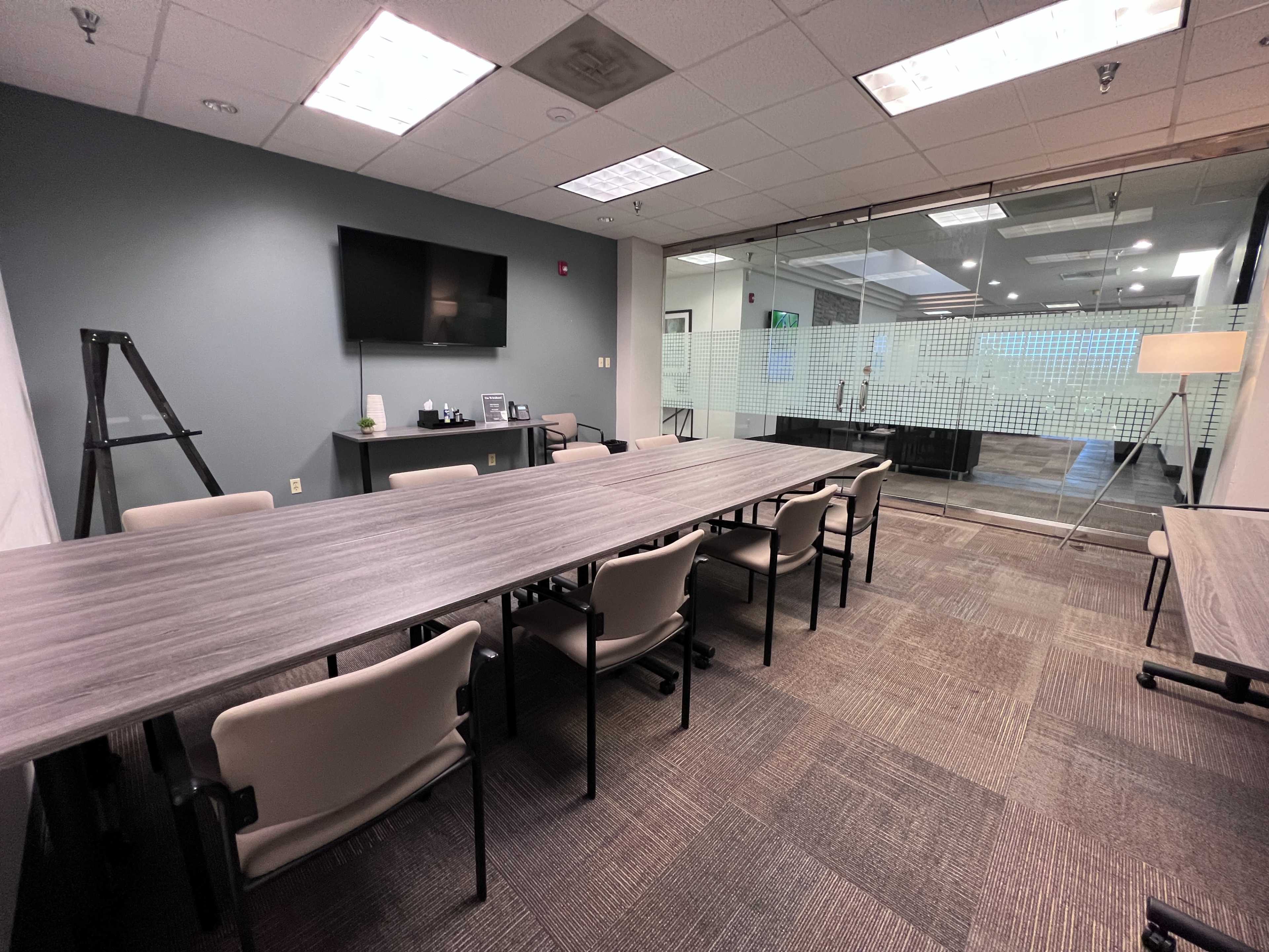 A meeting room with a long table, several chairs, a television, and glass walls leading to another area.