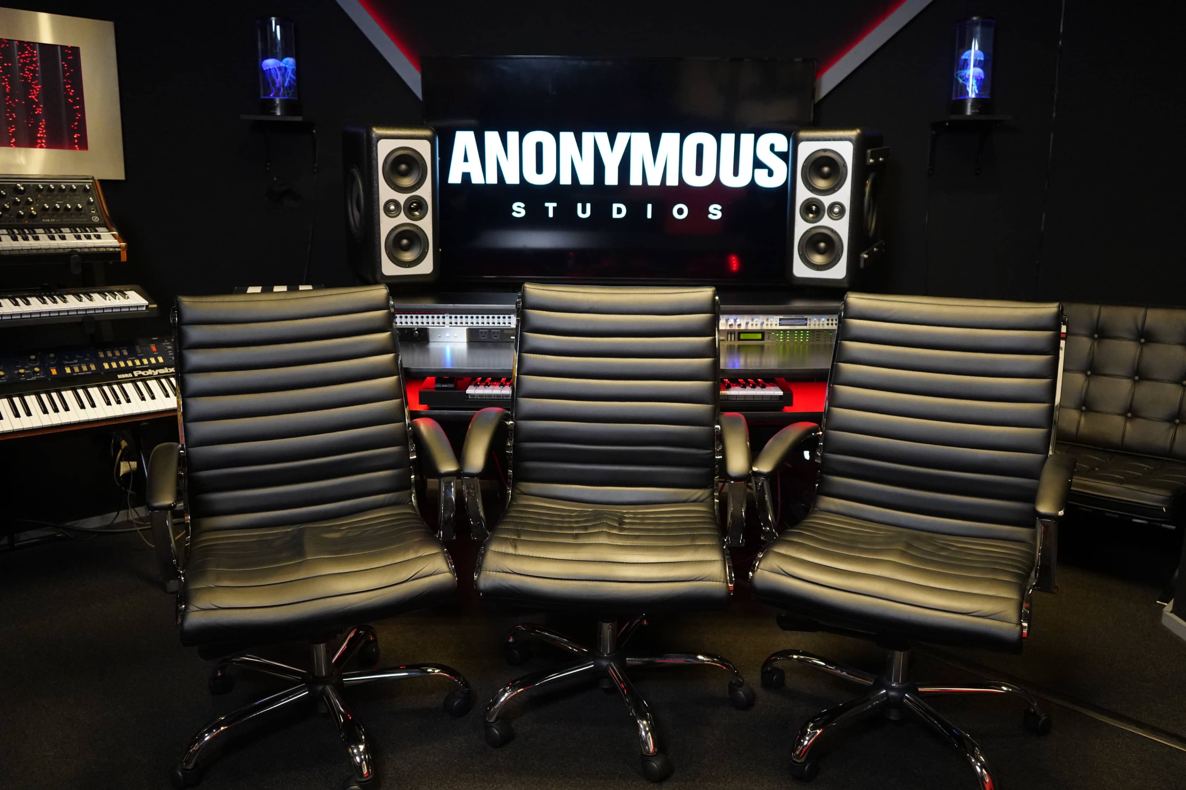 The image shows three black rolling office chairs in front of a sound studio setup with electronic equipment and a large screen displaying "ANONYMOUS STUDIOS."