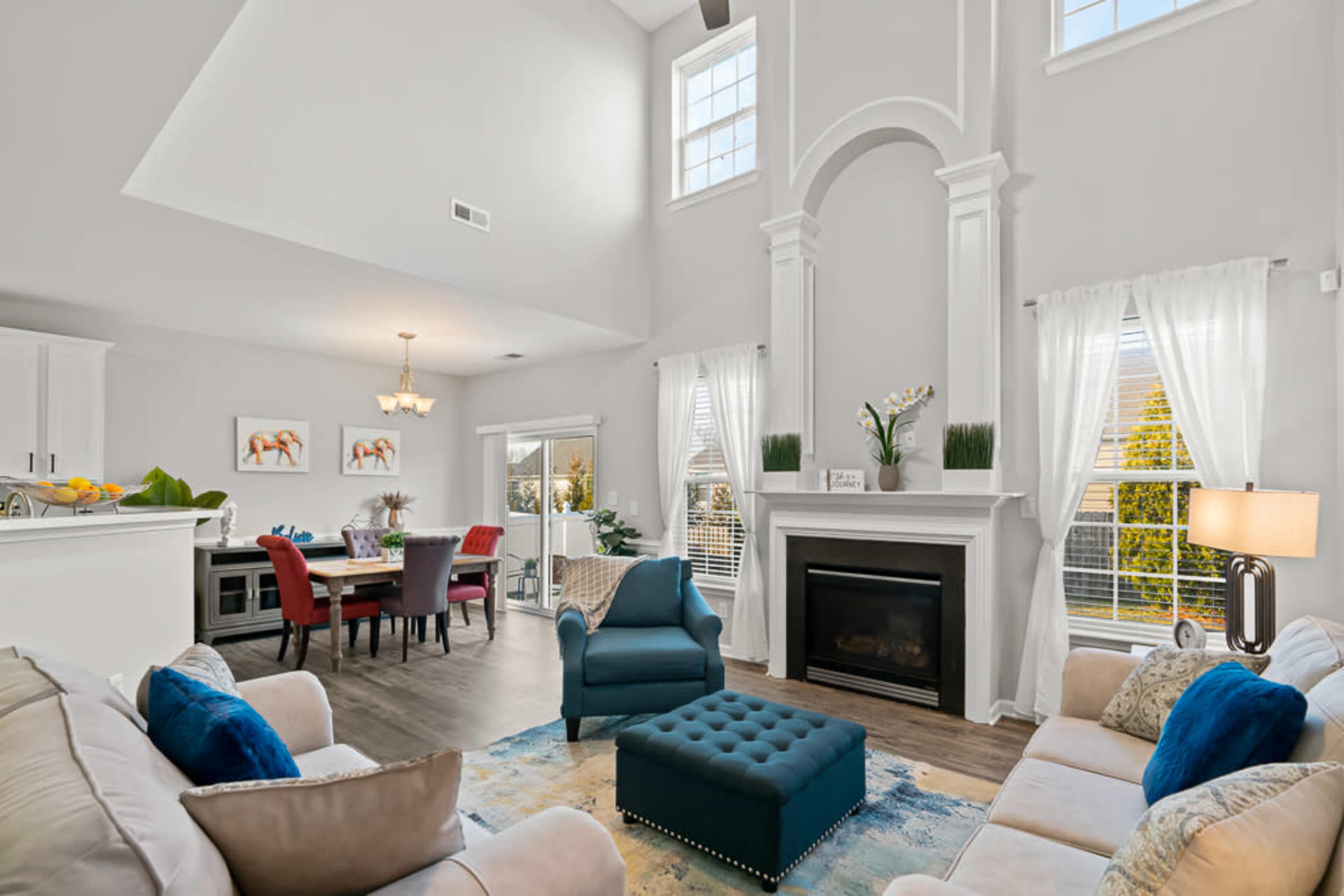 The living room features a vaulted ceiling, a fireplace with decorative molding, a blue tufted ottoman, and large windows allowing natural light.