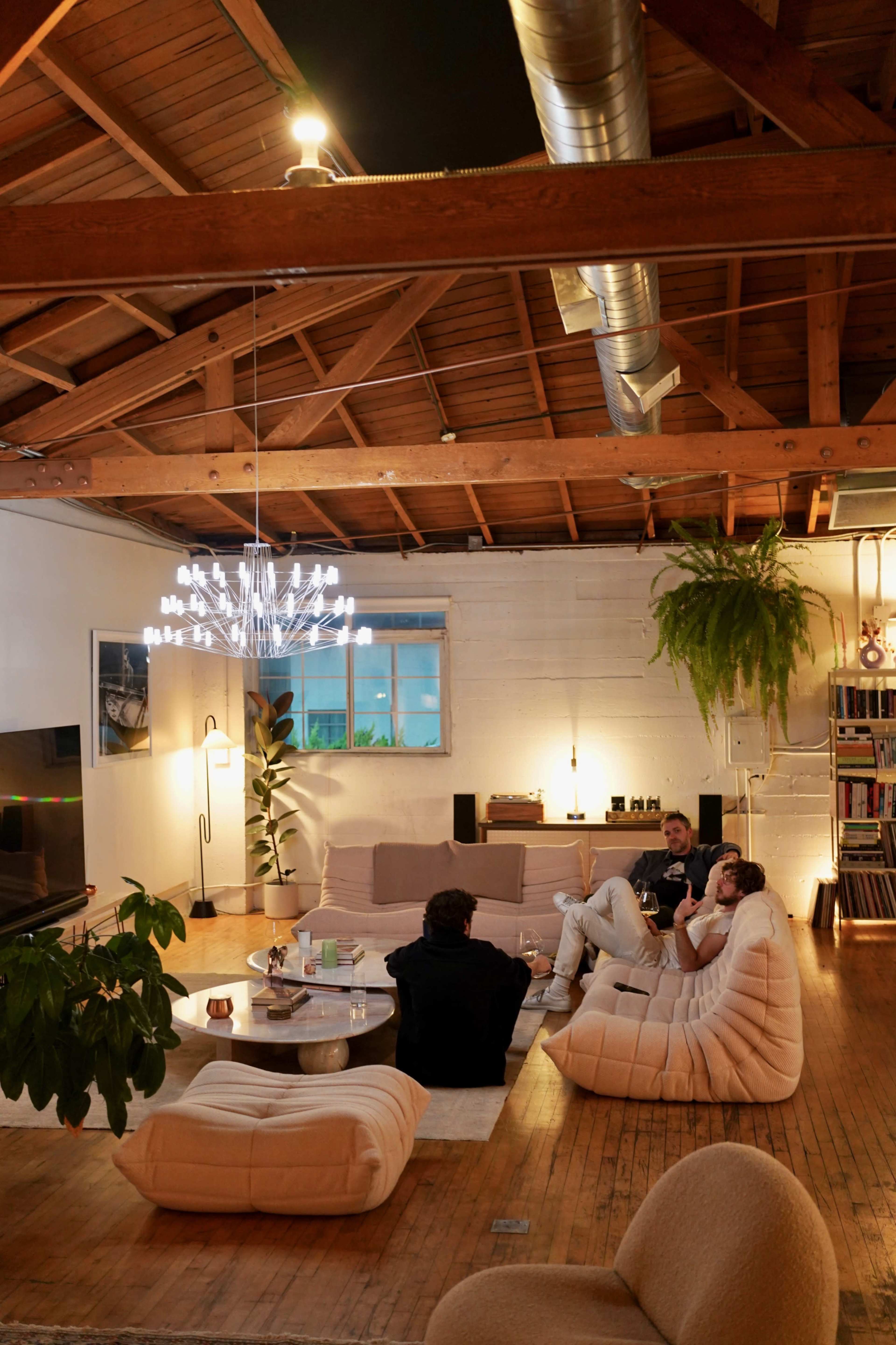 A spacious, modern living room with high wooden ceilings, a chandelier, and three people sitting on large, soft chairs engaged in conversation.