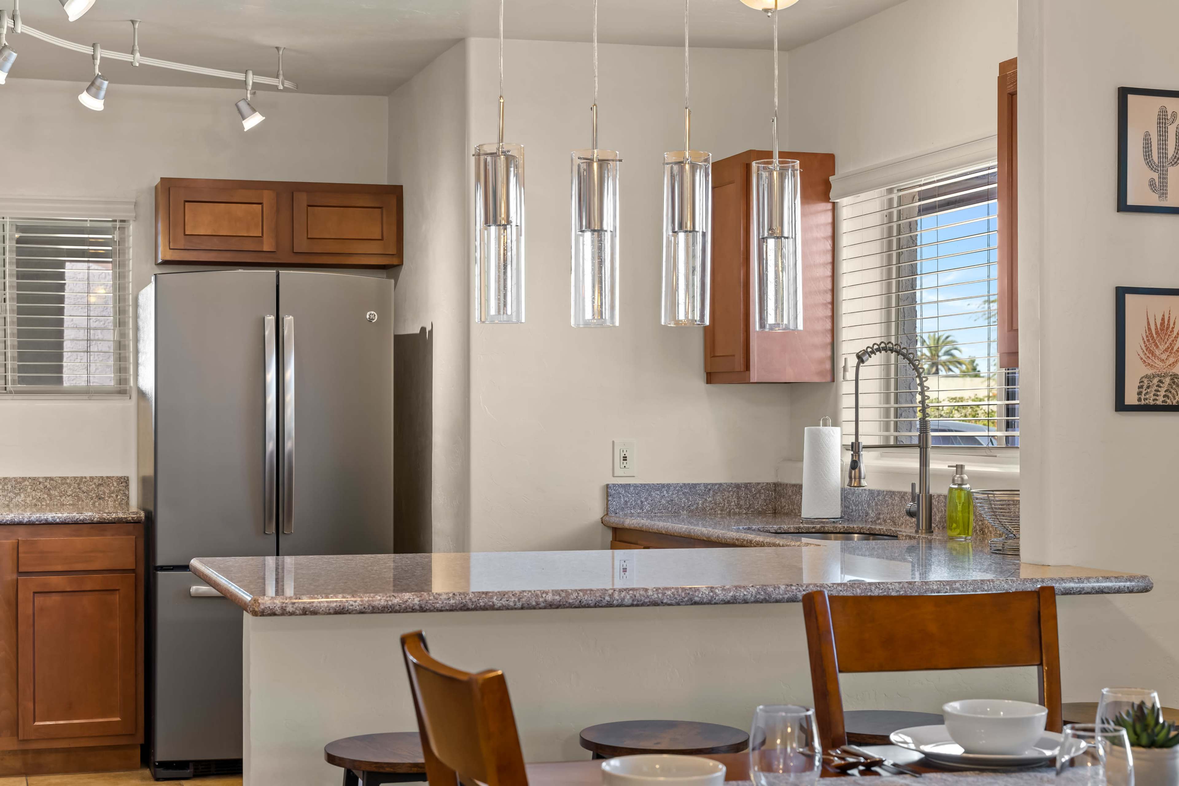 The image shows a modern kitchen with a gray refrigerator, wooden cabinets, a granite countertop, and pendant lighting above the kitchen island.