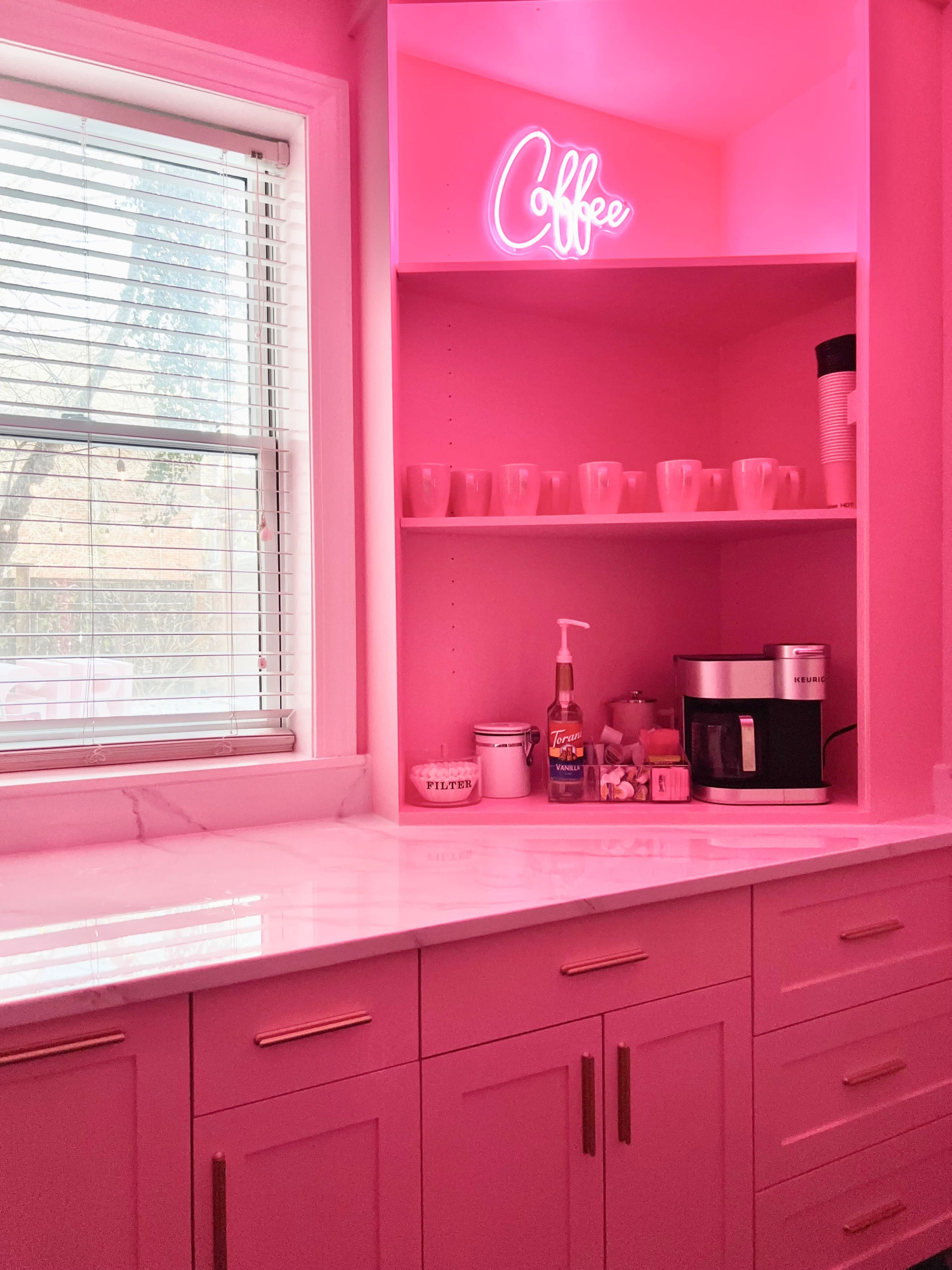 A brightly lit kitchen features pink cabinetry, a coffee maker, and a neon sign that says "Coffee."