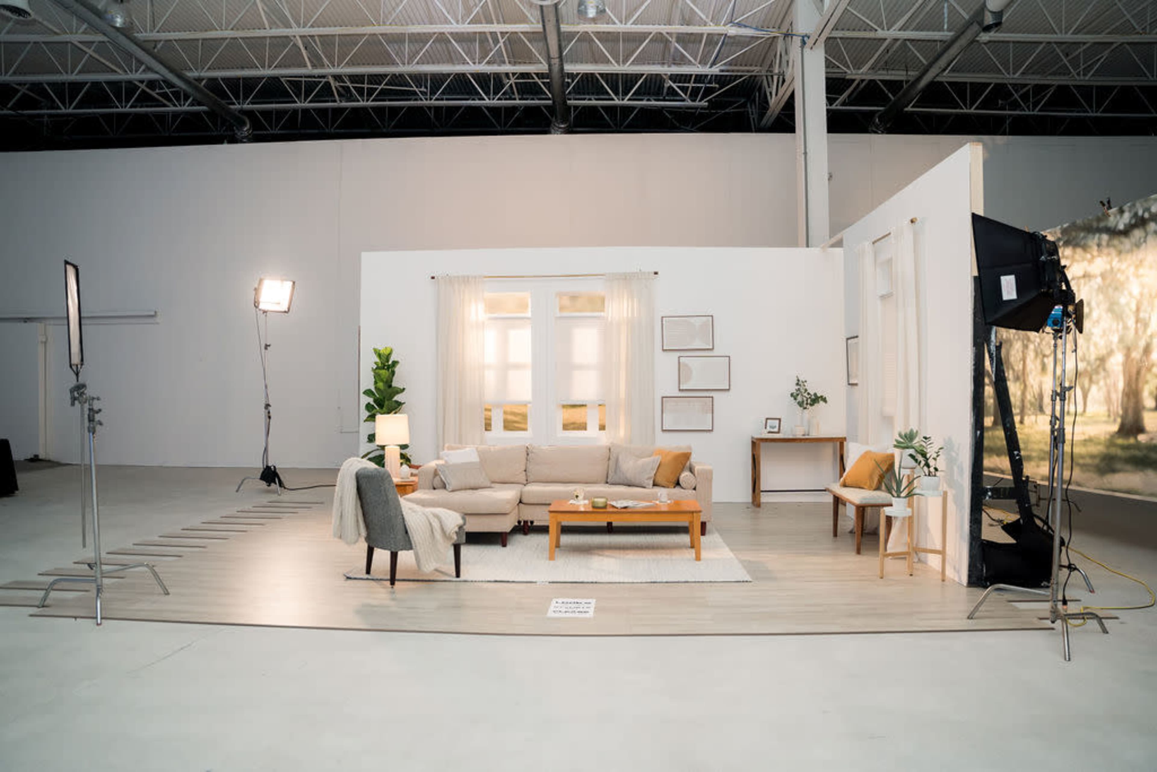 The image shows a staged living room setup with a sofa, coffee table, and decorative plants in a large, well-lit studio space.