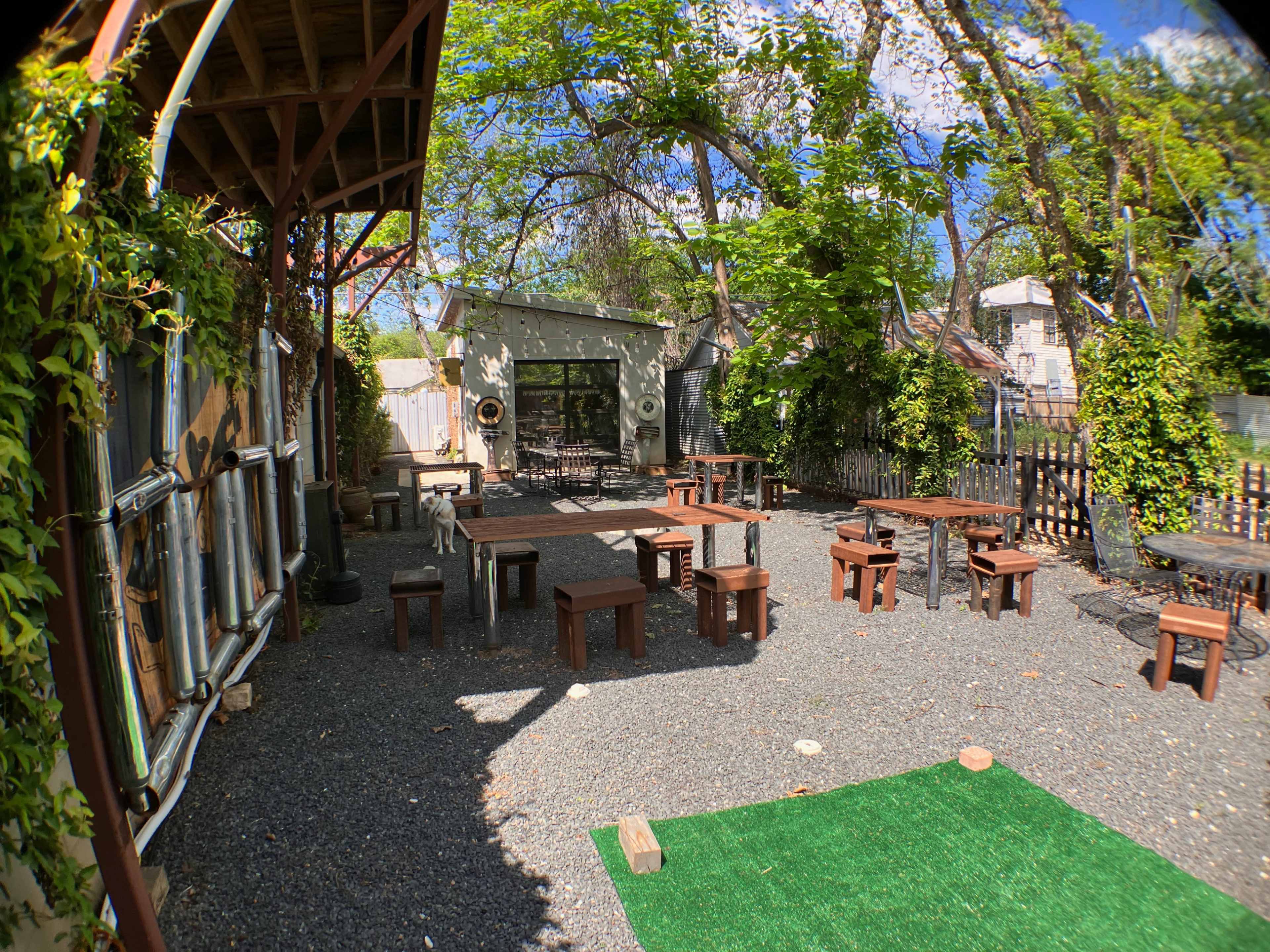 The image shows an outdoor seating area with wooden benches arranged around a gravel surface, surrounded by greenery and a building in the background.