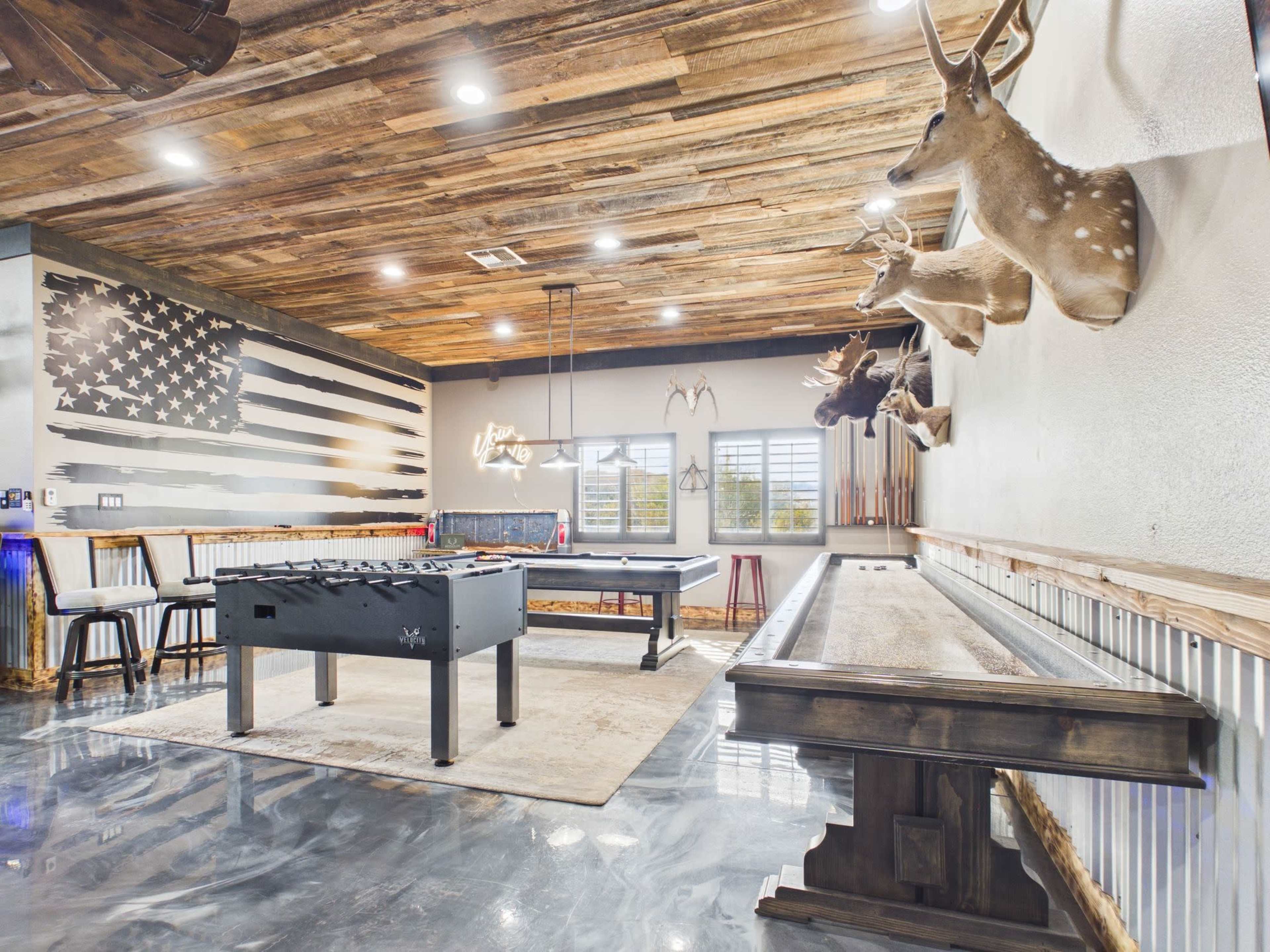 The image shows a rustic game room featuring a foosball table, a shuffleboard table, mounted deer heads on the wall, and a large American flag mural.