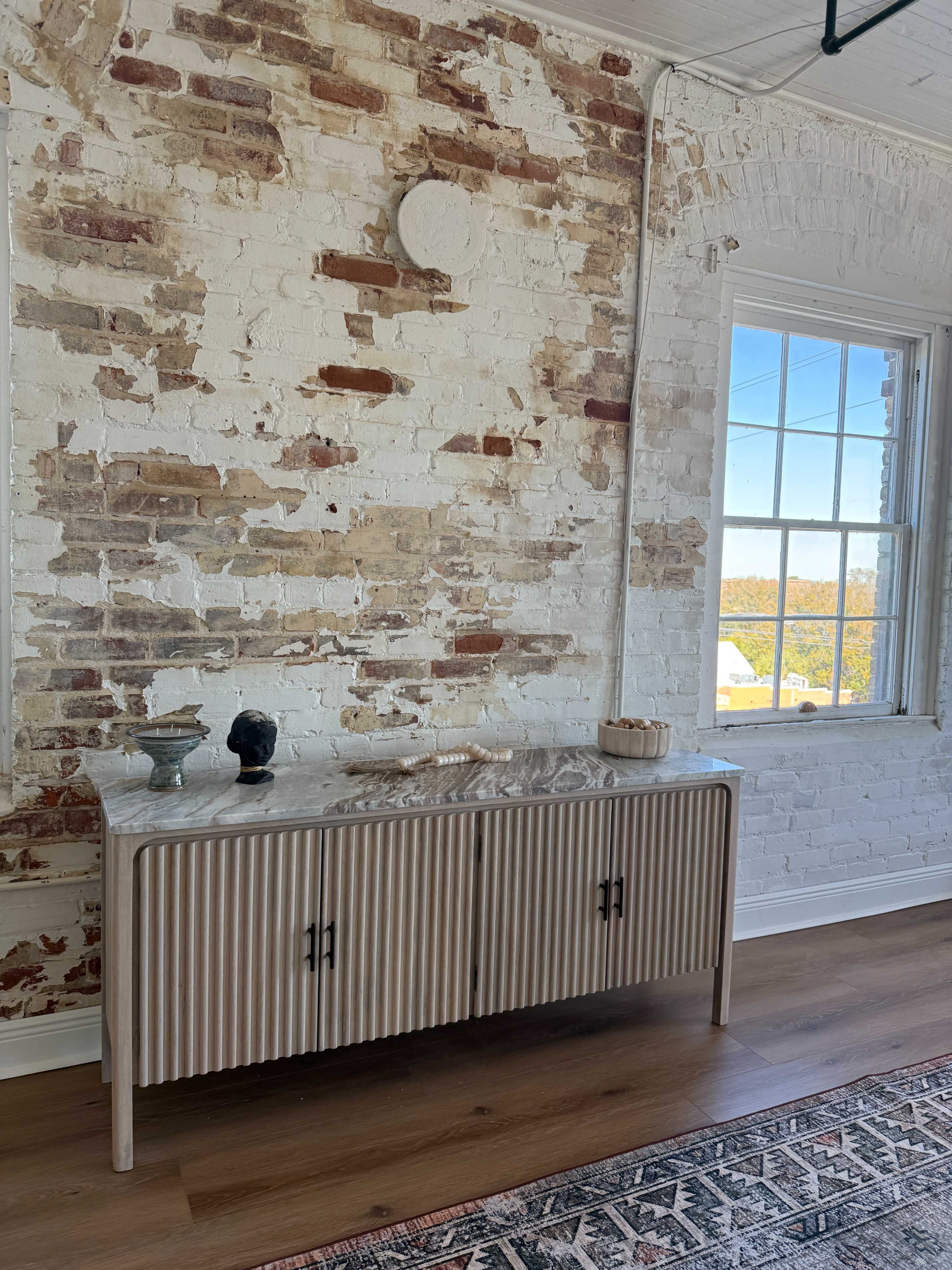 The image features a wooden sideboard with ribbed detailing placed against a wall of exposed brick, with a large window providing natural light.