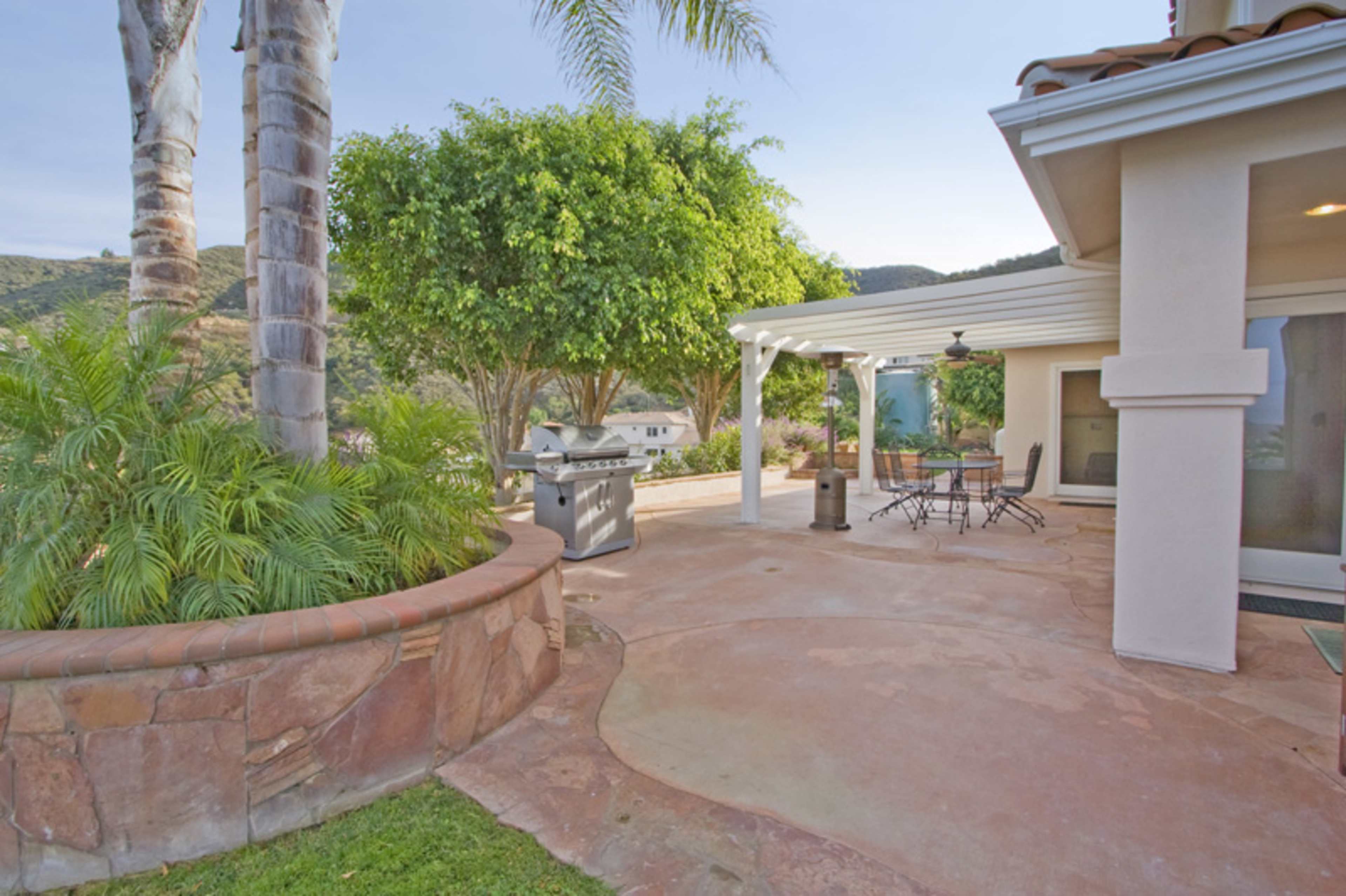 A patio area with a grill, table, and chairs surrounded by greenery and hills in the background.
