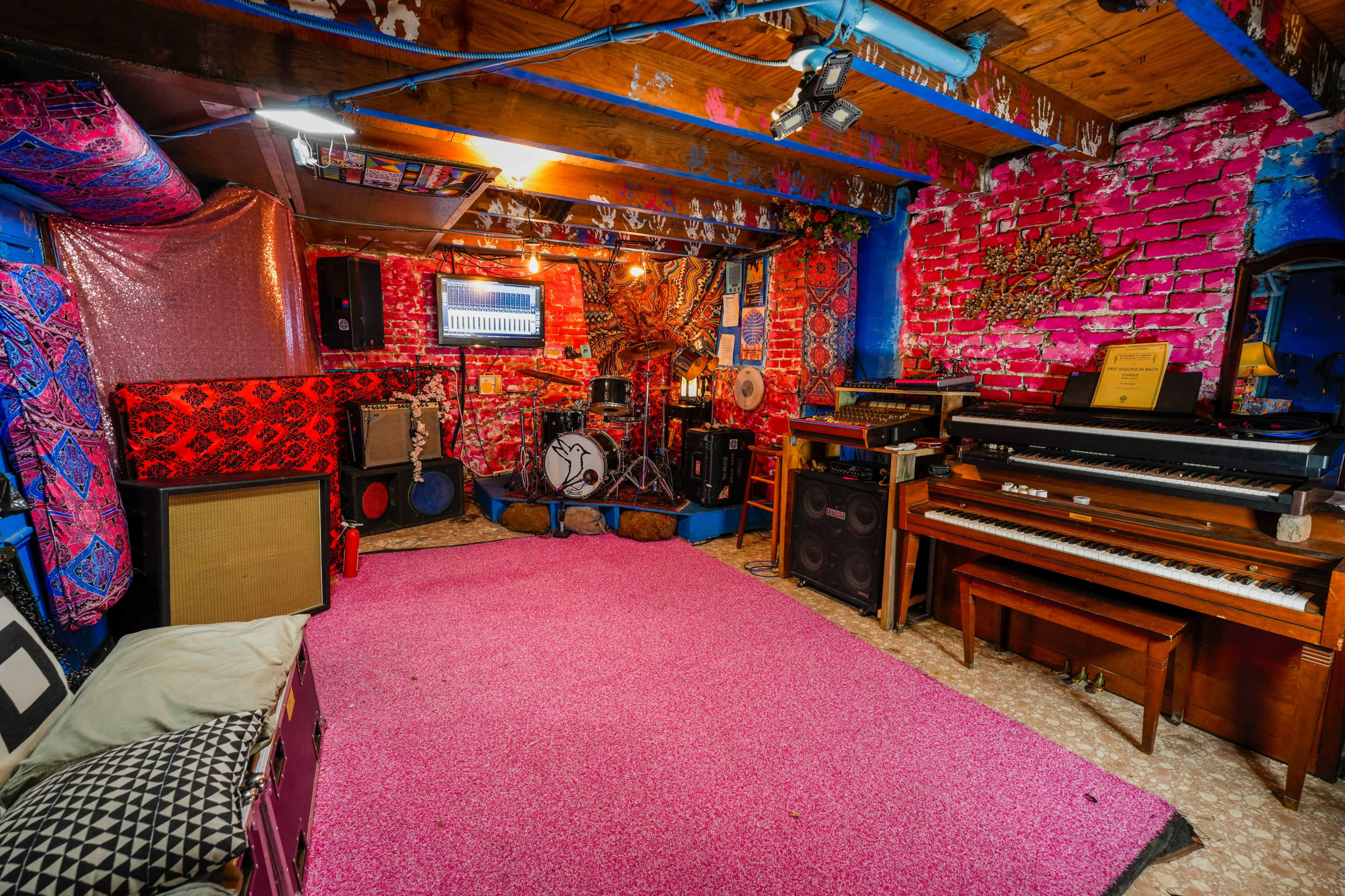 The image shows a colorful basement room featuring a drum set, a piano, and various amplifiers, all arranged around a pink carpet.